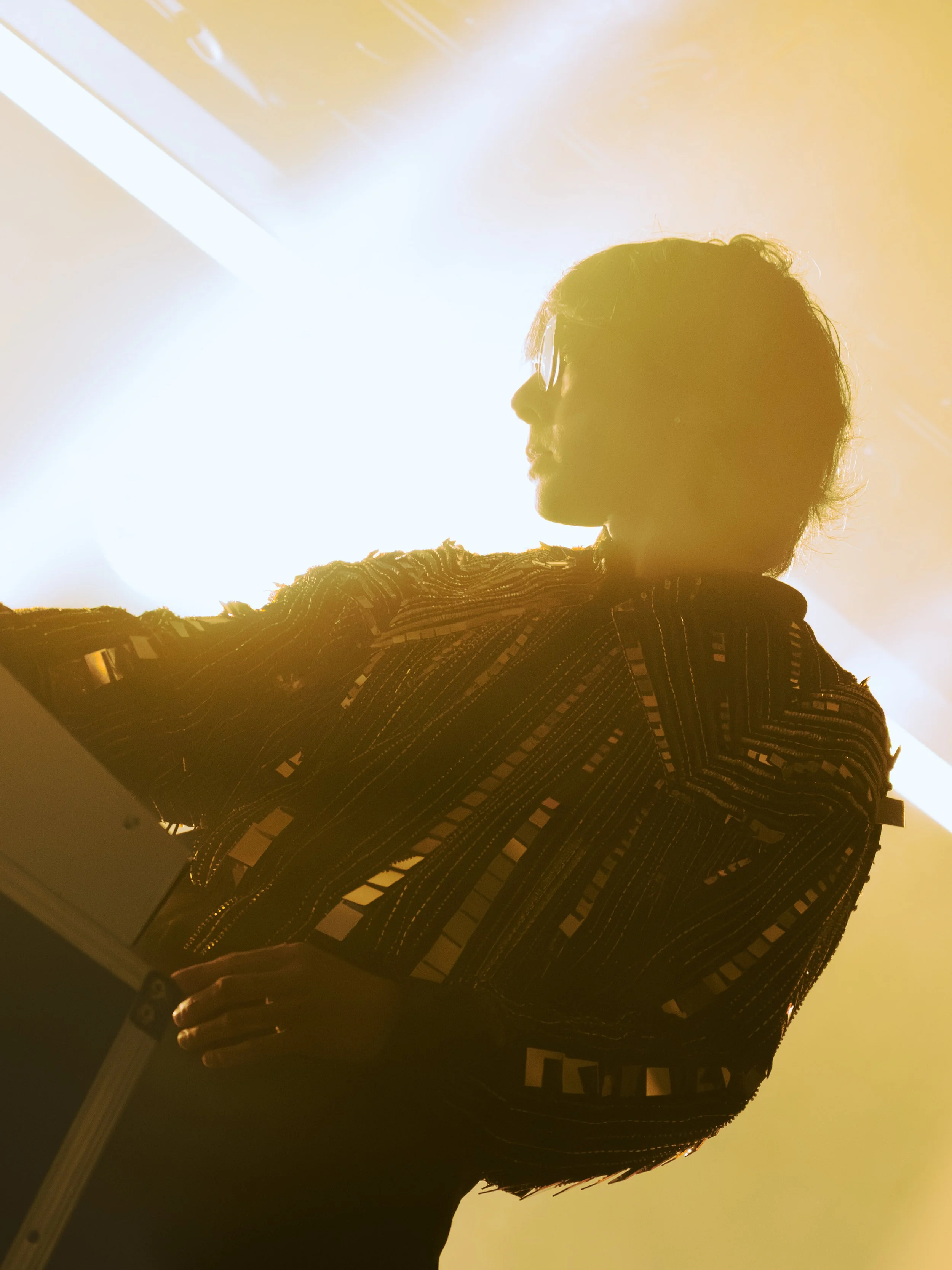 Silhouette of a person with short hair and glasses looking to the side, standing in front of bright sunlight, wearing a patterned jacket.