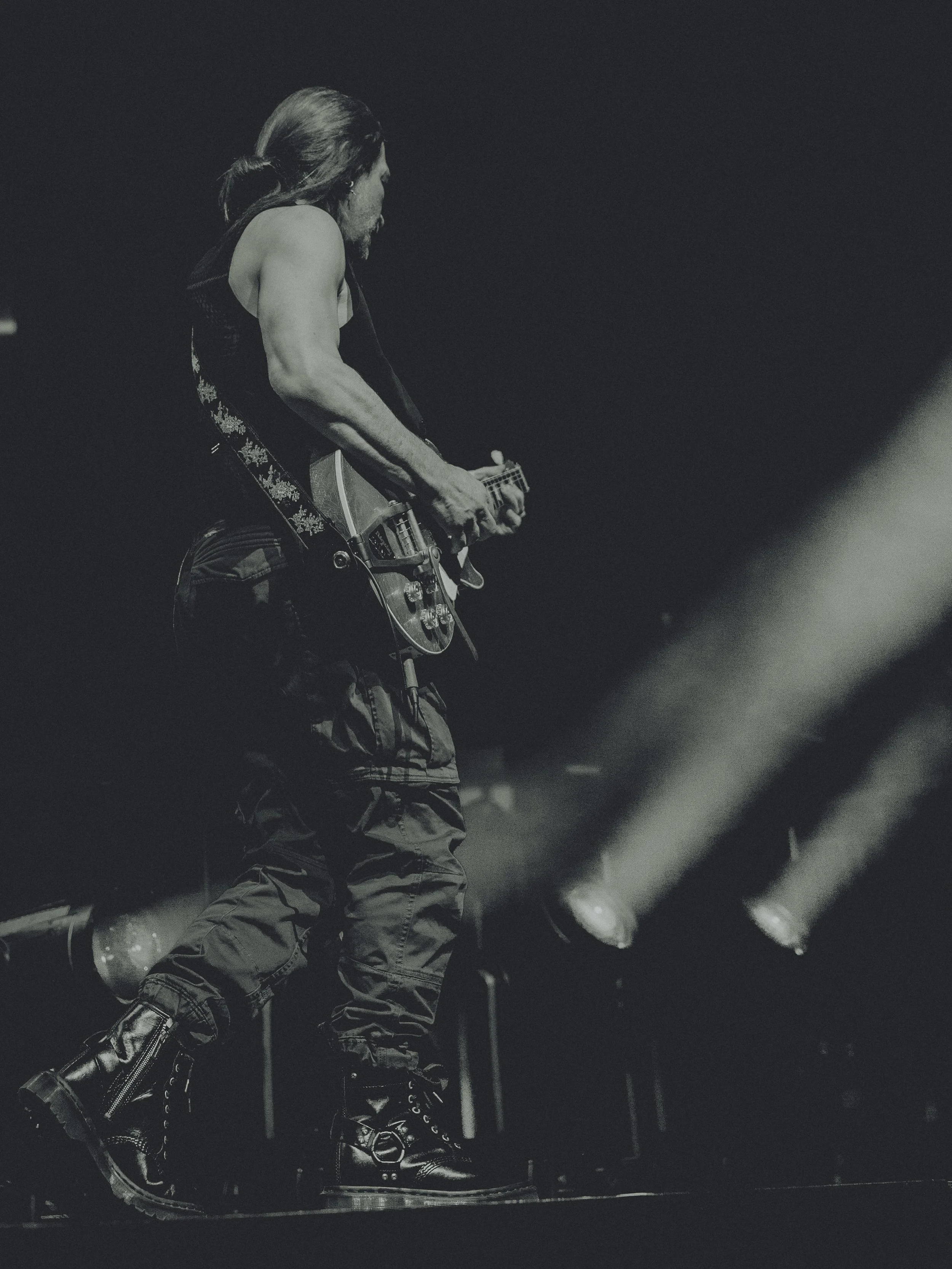 A black and white photo of a male guitarist performing on stage, with long hair tied back, wearing a sleeveless shirt, cargo pants, and boots. Stage lights shine in the background.