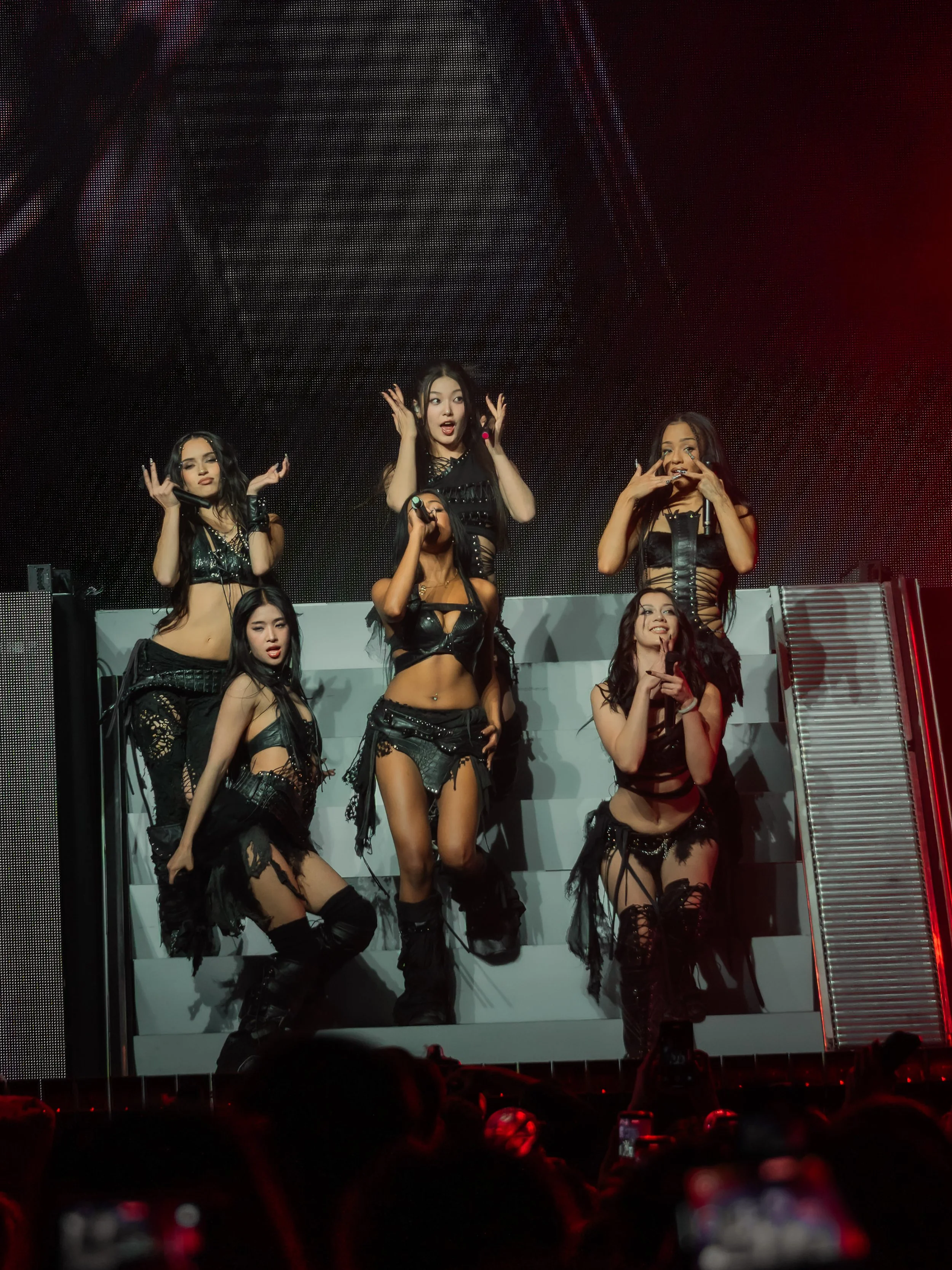 Group of six female performers dressed in black stage outfits performing on a platform during a concert.