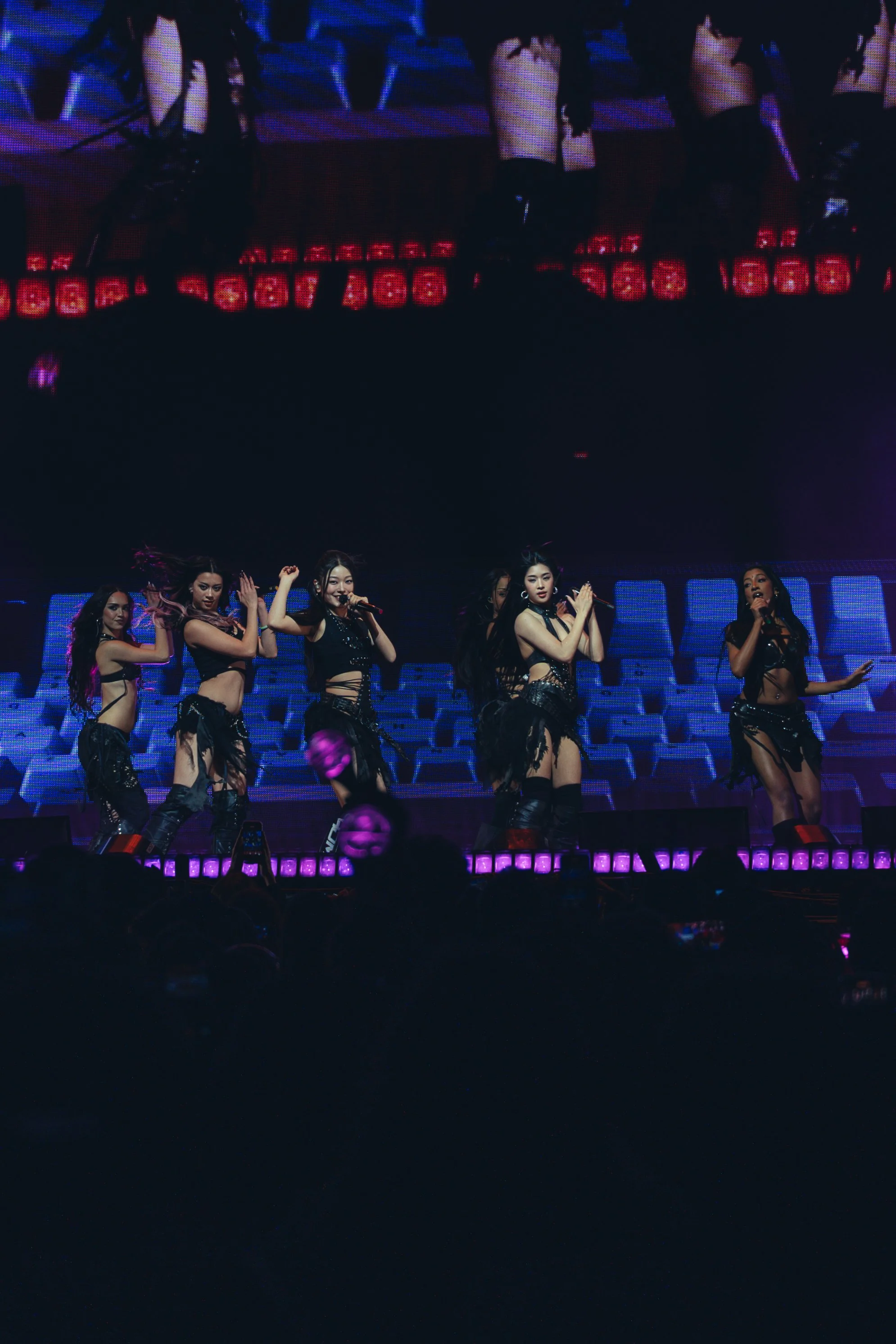 A group of female performers on a stage, singing and dancing in black outfits, with an audience watching.