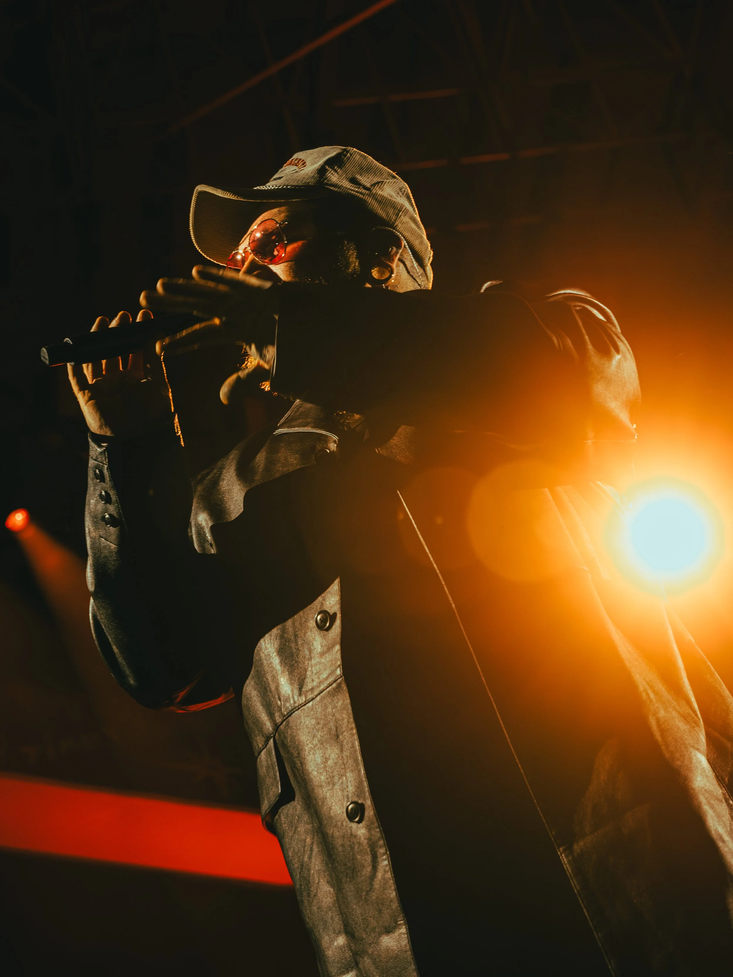 Performer wearing a cap and sunglasses, holding a microphone, illuminated by stage light in a dark setting.