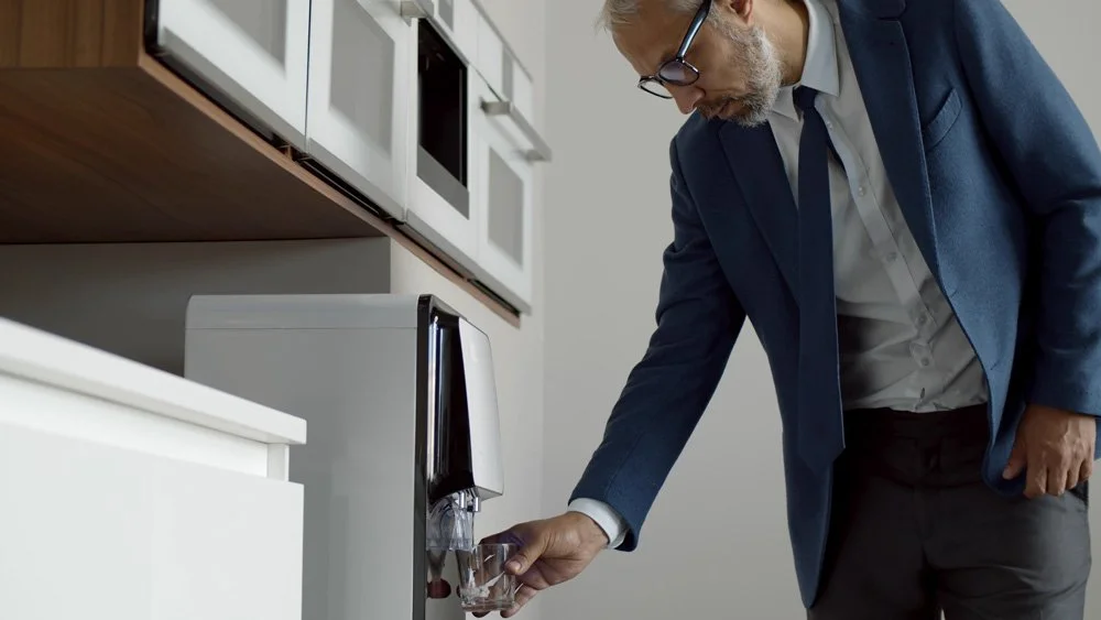 A man in business attire filling a glass with water from a water cooler in an office kitchen.