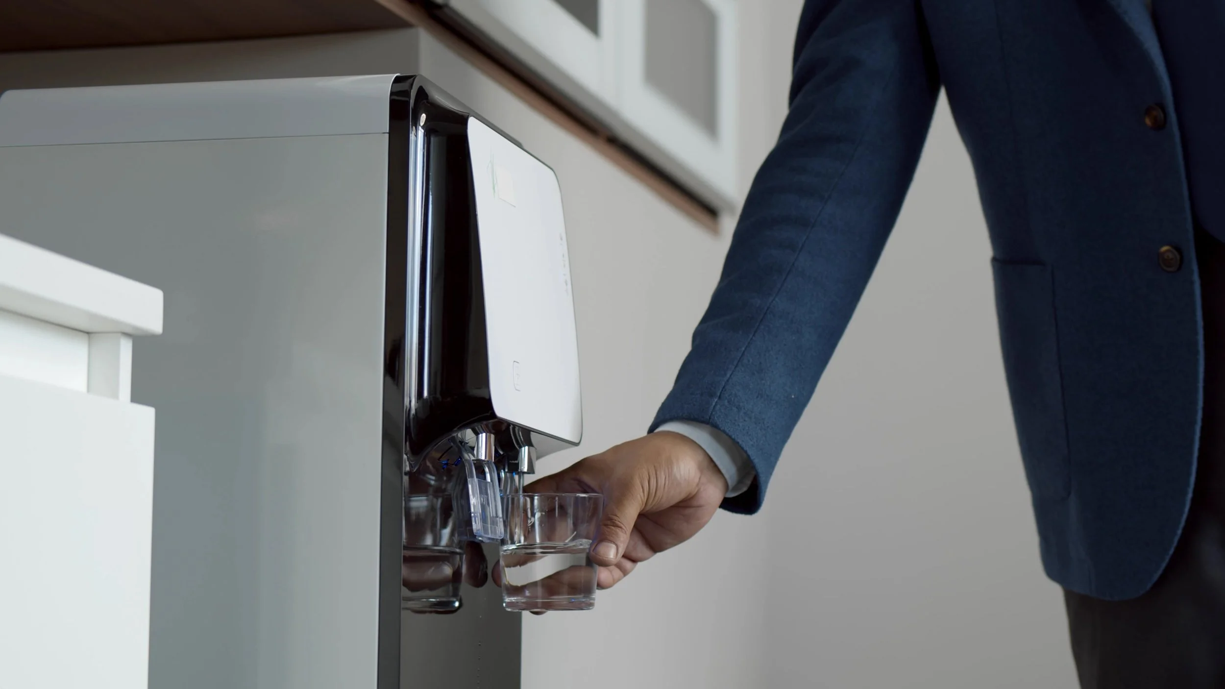 Person in a blue blazer filling a drinking glass with water from a water dispenser.