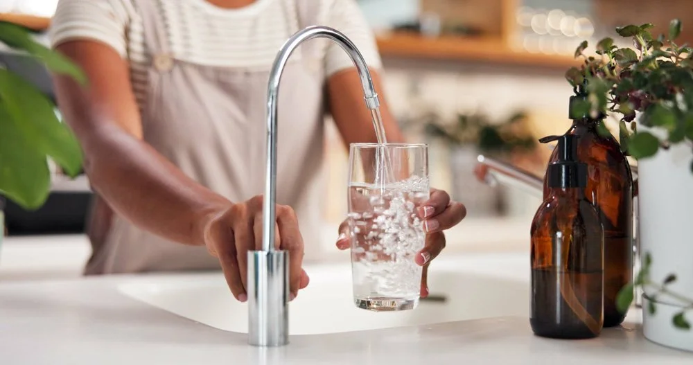 Person filling a glass with water at a kitchen sink with plants nearby.