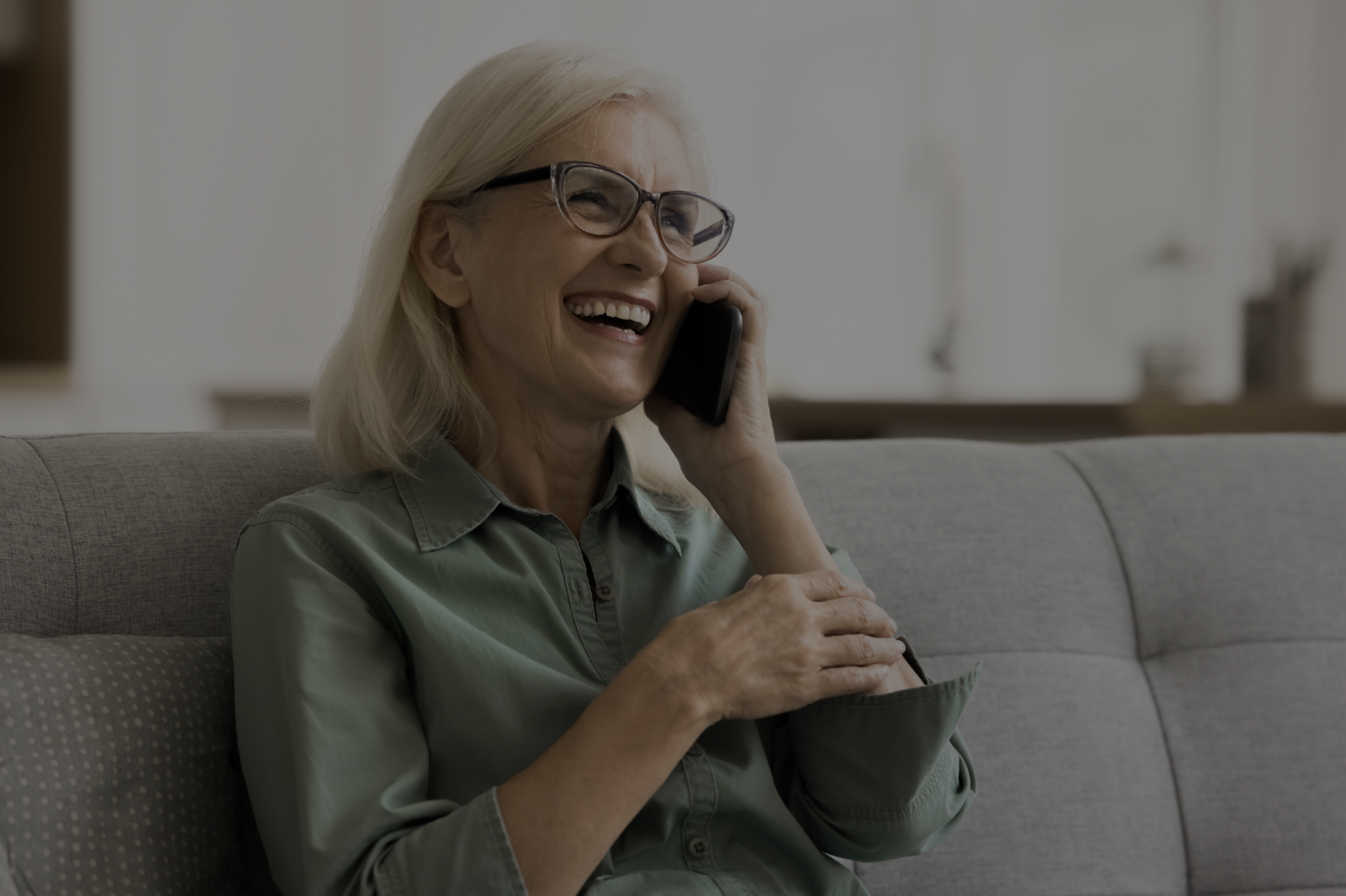 An elderly woman with white hair, glasses, and a green blouse, smiling while talking on a black phone in a cozy living room.