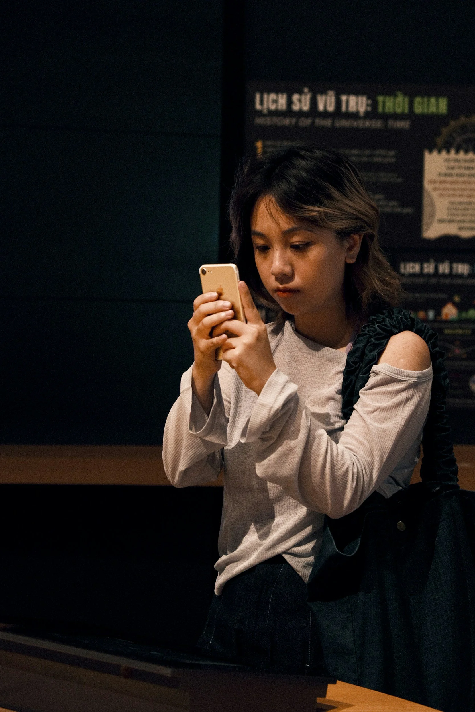 A young woman with wavy hair is looking at her smartphone while standing indoors. She is wearing a gray long-sleeve shirt with a shoulder cut-out and carrying a black bag over her shoulder. In the background, there is a dark wall with a poster that has some Vietnamese text.