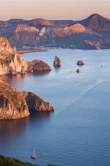 Paesaggio costiero con scogliere rocciose, mare calmo e montagne sullo sfondo al tramonto.