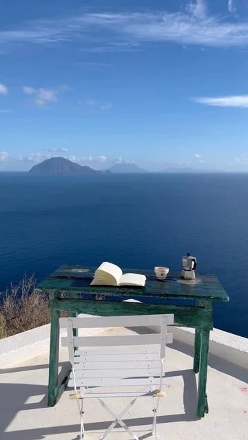 Mare aperto con isole lontane, cielo sereno con alcune nuvole, tavolo con libro, tazza e macchina da caffè, posizione vista panoramica.