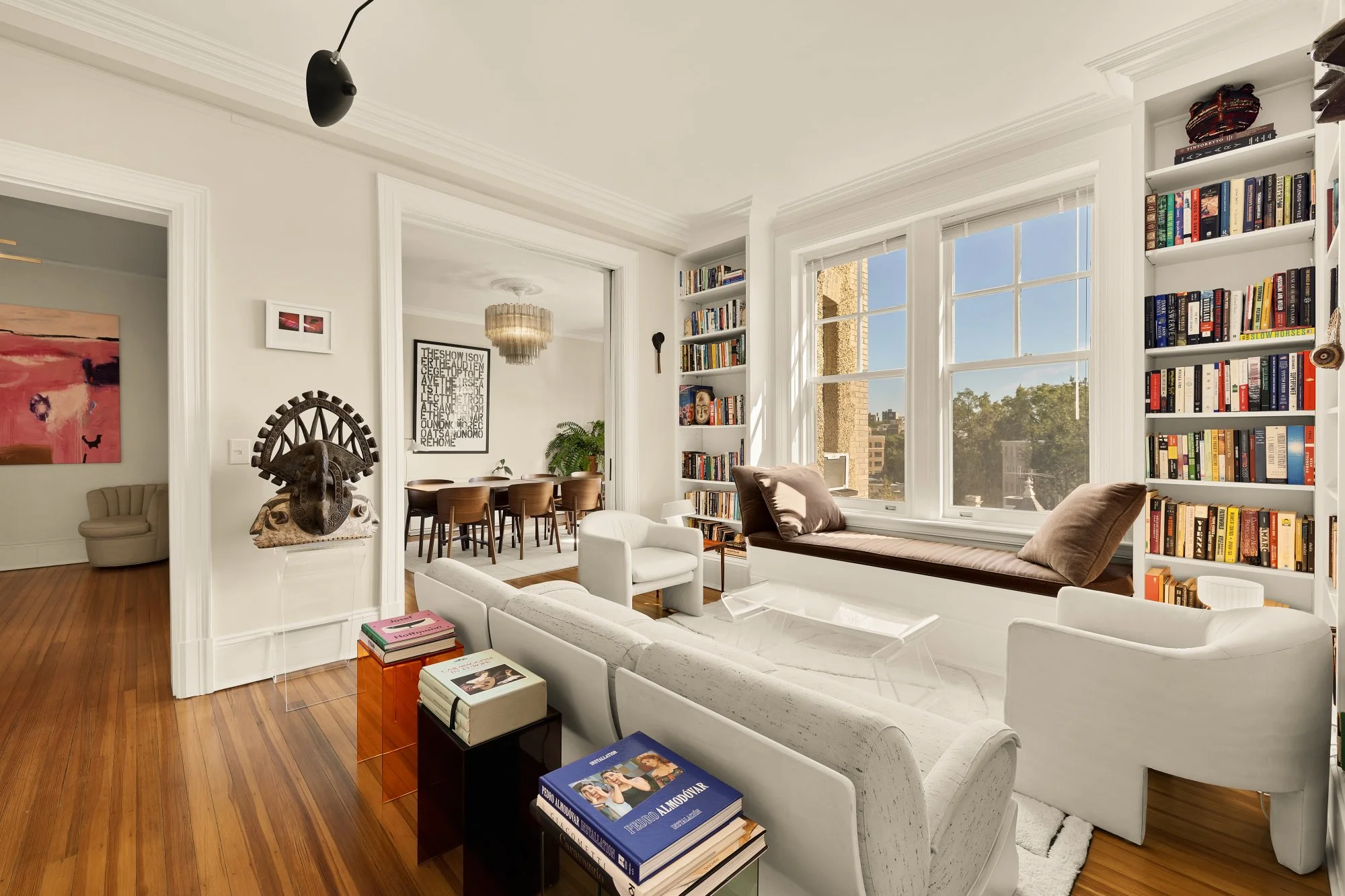 Sunlit living room with white walls, large window seats, bookshelves filled with books, a white sofa, armchairs, and a glass coffee table. Visible dining area with a chandelier and a wall art in the background.