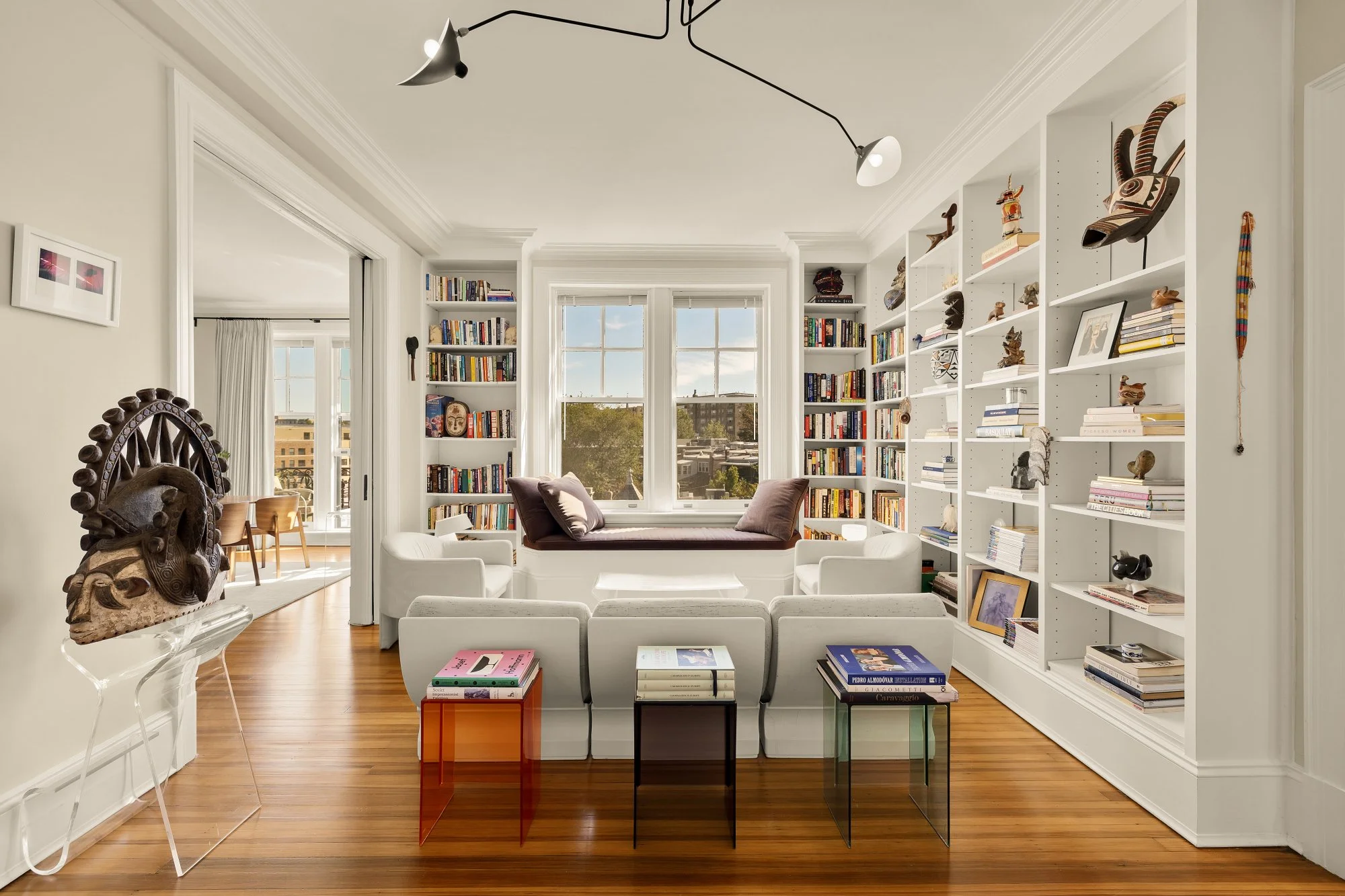 A bright living room with large windows, white built-in bookshelves filled with books and art, white seating, and wooden flooring. There is a window seat with pillows, a small set of colorful side tables, and a sculpture on a stand.