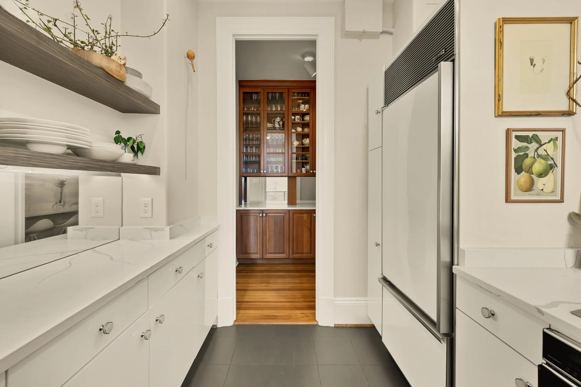 A modern kitchen with white cabinets, a marble countertop, open wooden shelves with plates and bowls, and decorative art on the wall. A doorway leads to a room with wooden floors and a glass-fronted cabinet.