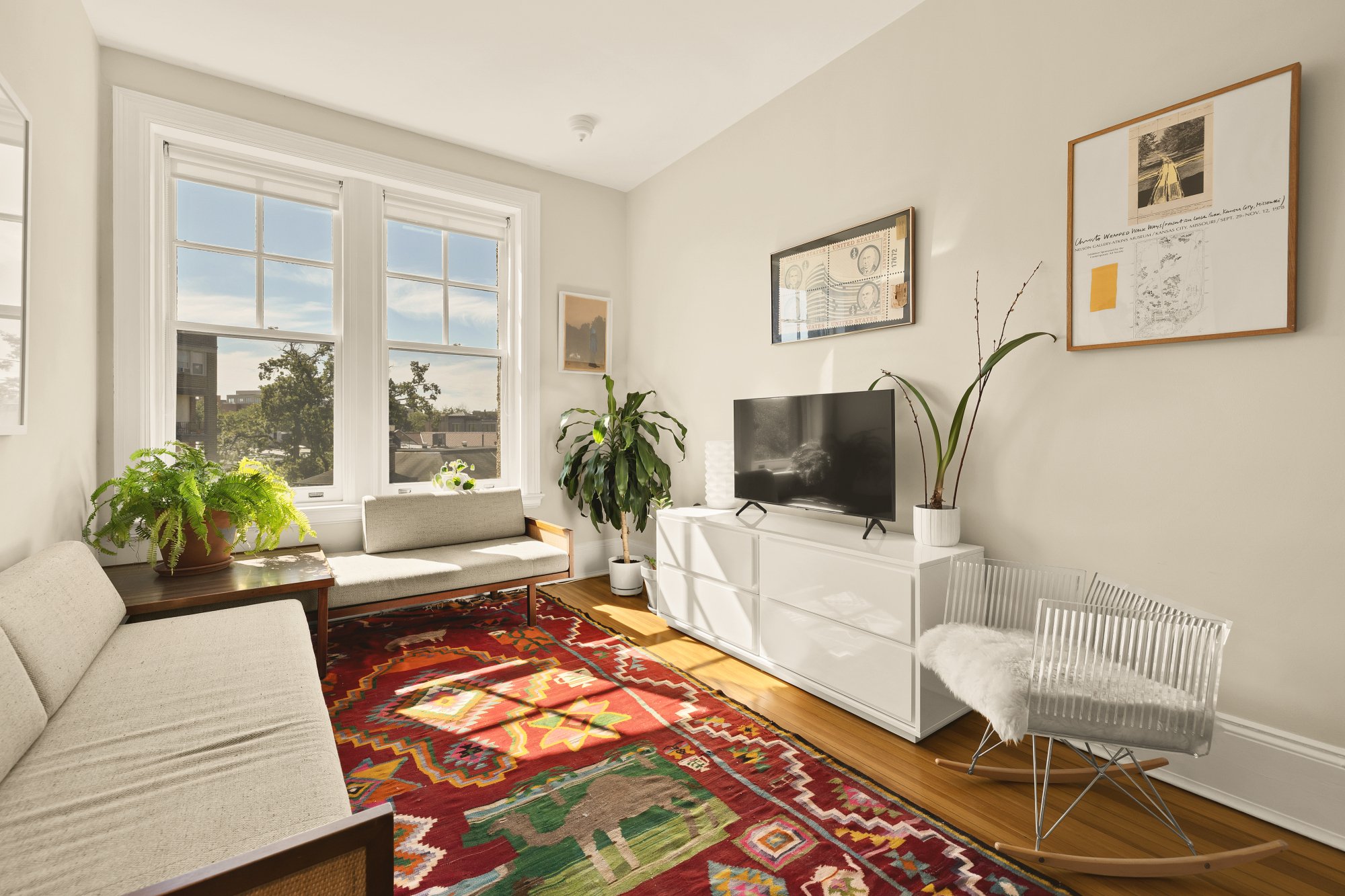 Bright living room with large windows, a colorful patterned rug, a white TV stand, a flatscreen TV, green plants, framed artwork, a beige sofa, a white rocking chair with fur, and natural light streaming in.
