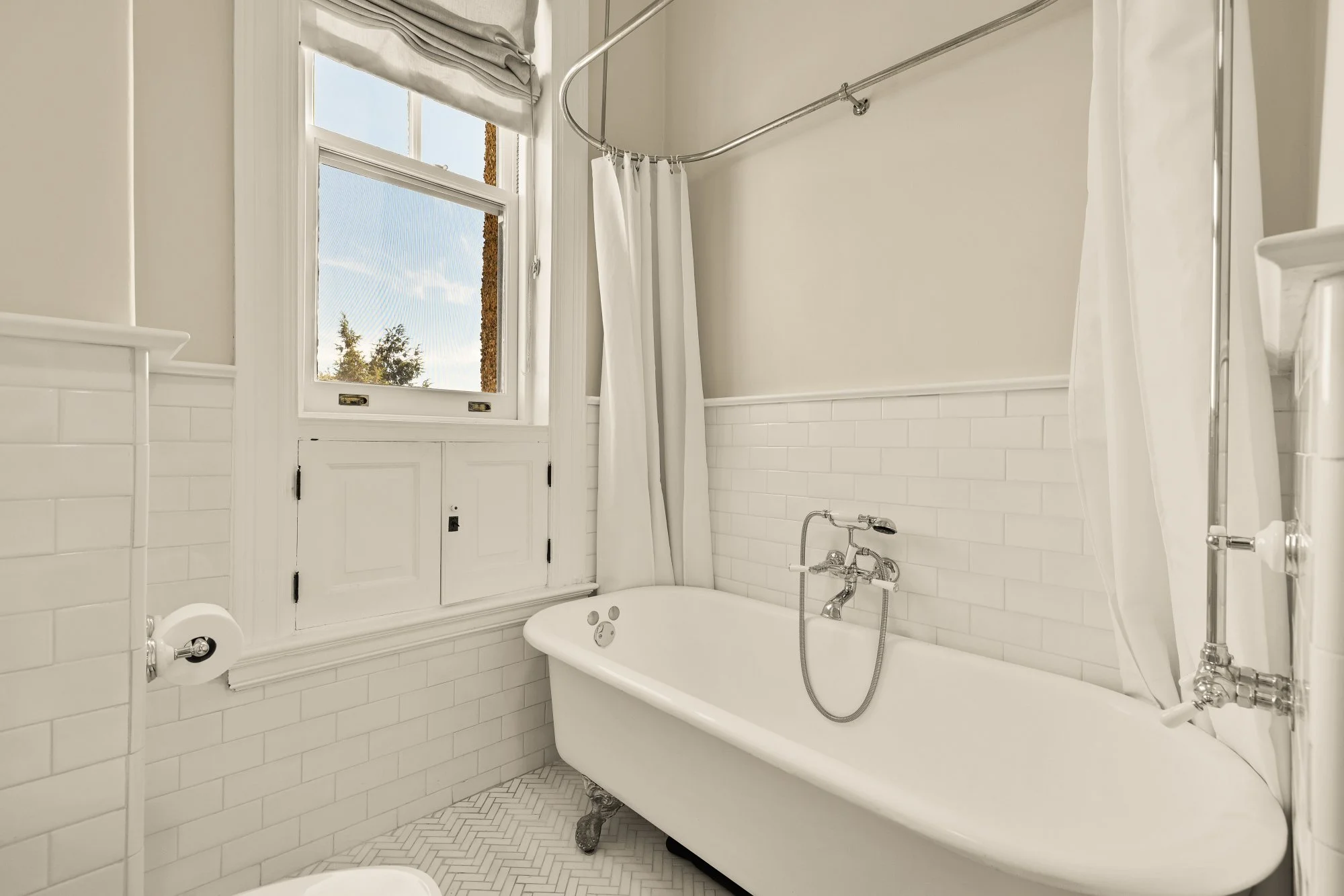 Bathroom with a clawfoot bathtub, window with a view of trees, white tiled walls, and curtains.