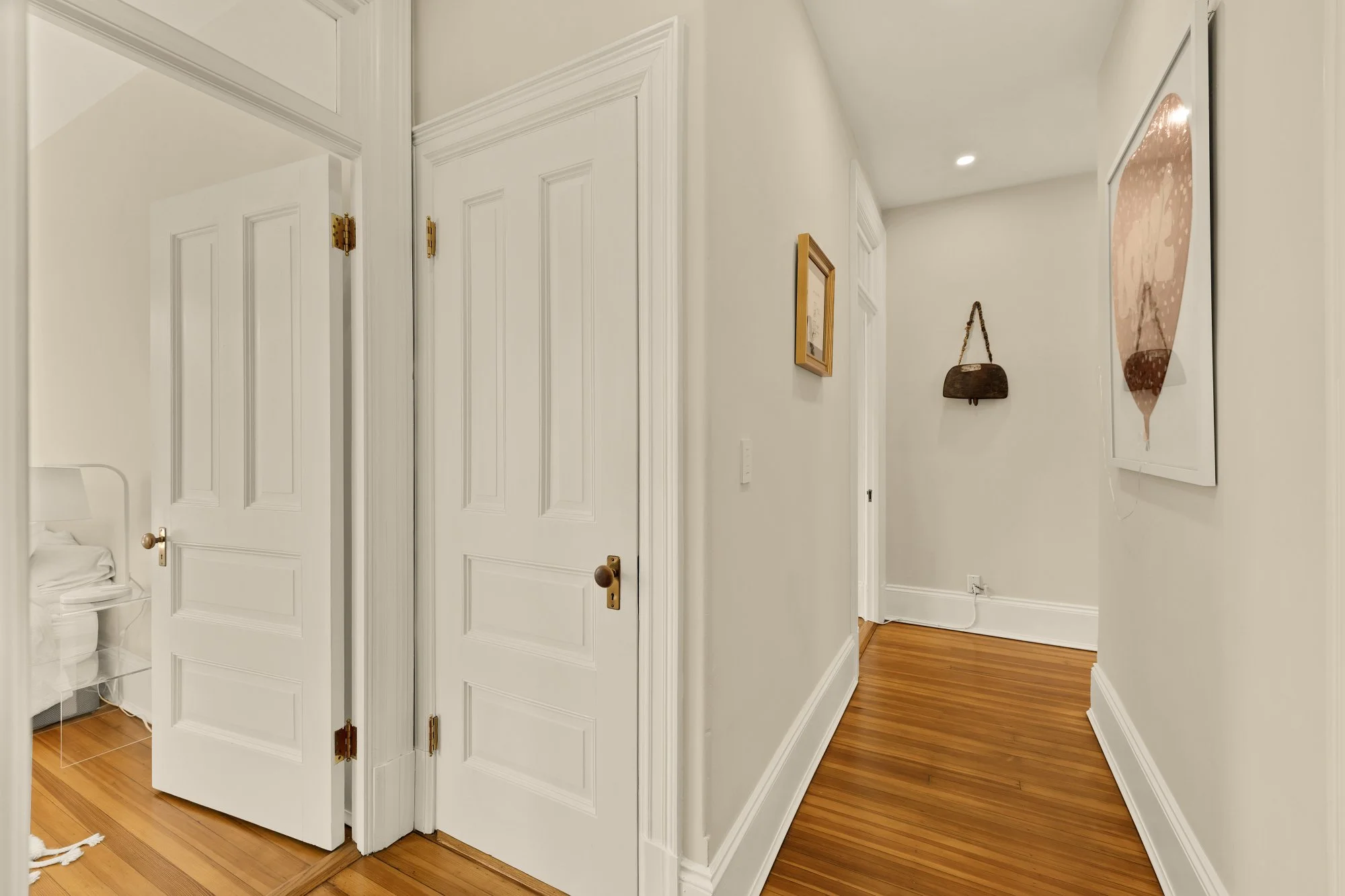 Hallway with white walls, wooden floors, and decorative molding. Doors along the hallway with brass hinges and handles, one door slightly open revealing a bedroom. Artwork on the walls, including a framed picture and a large abstract piece. A bag han