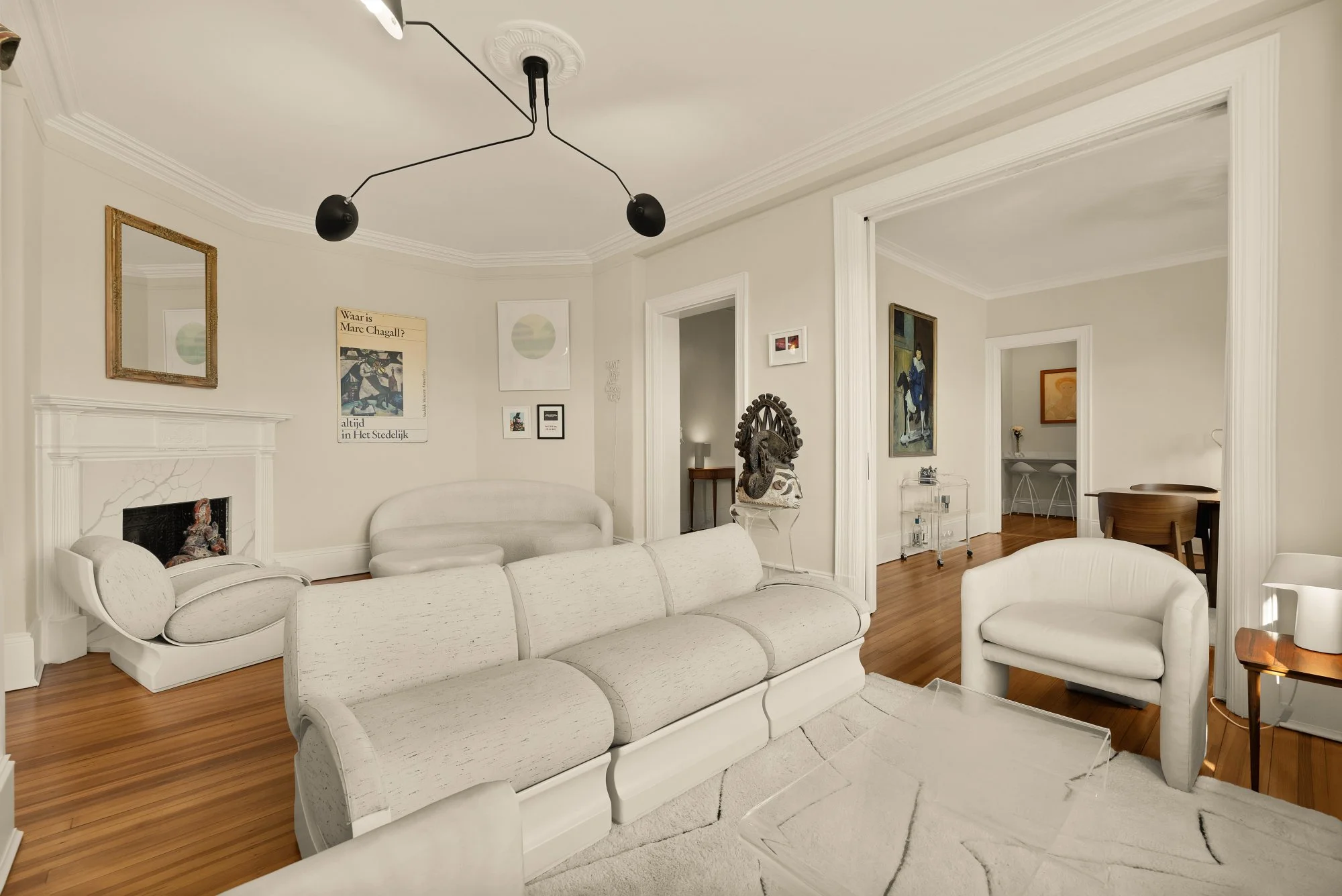 Living room with white sofa, armchair, modern black ceiling light, artwork on walls, fireplace, wooden floors, and doorway to another room.