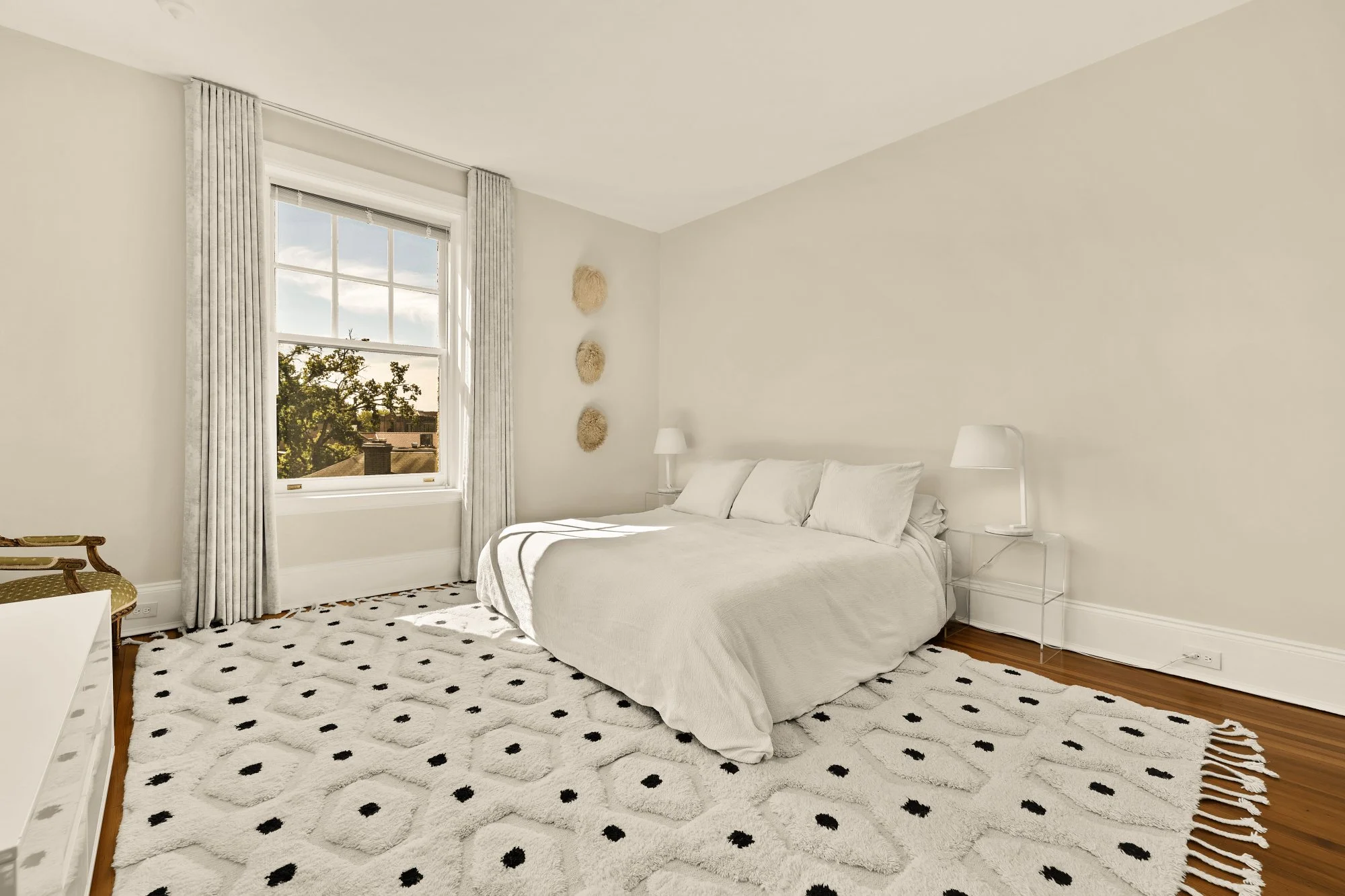 A bright bedroom with white walls, a large window with curtains, a bed with white bedding, a patterned rug, and small lamps on bedside tables.