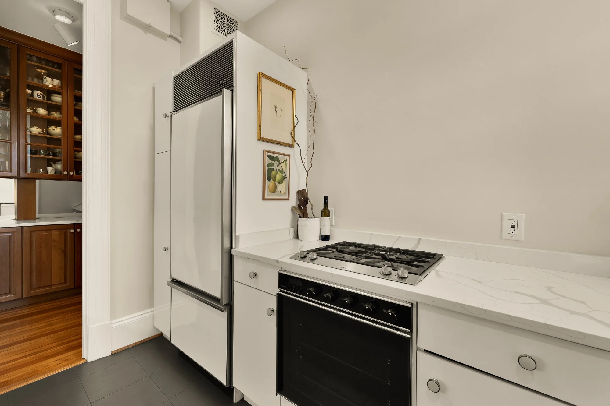 Kitchen corner with a white marble countertop, a black oven, and a white refrigerator. There are two framed pictures on the wall, and some kitchen utensils and a bottle of wine on the counter. Part of a wooden cabinet with dishes is visible in the ad