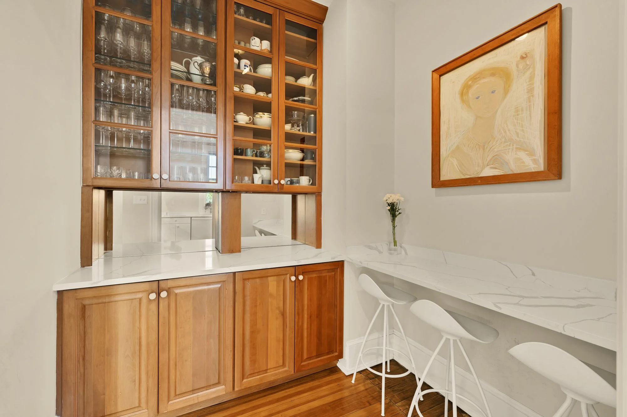 Kitchen corner with wooden cabinets filled with glassware and dishes, white marble countertop, white barstools, and a light-colored wall with a framed painting of a woman and a small vase of white flowers.