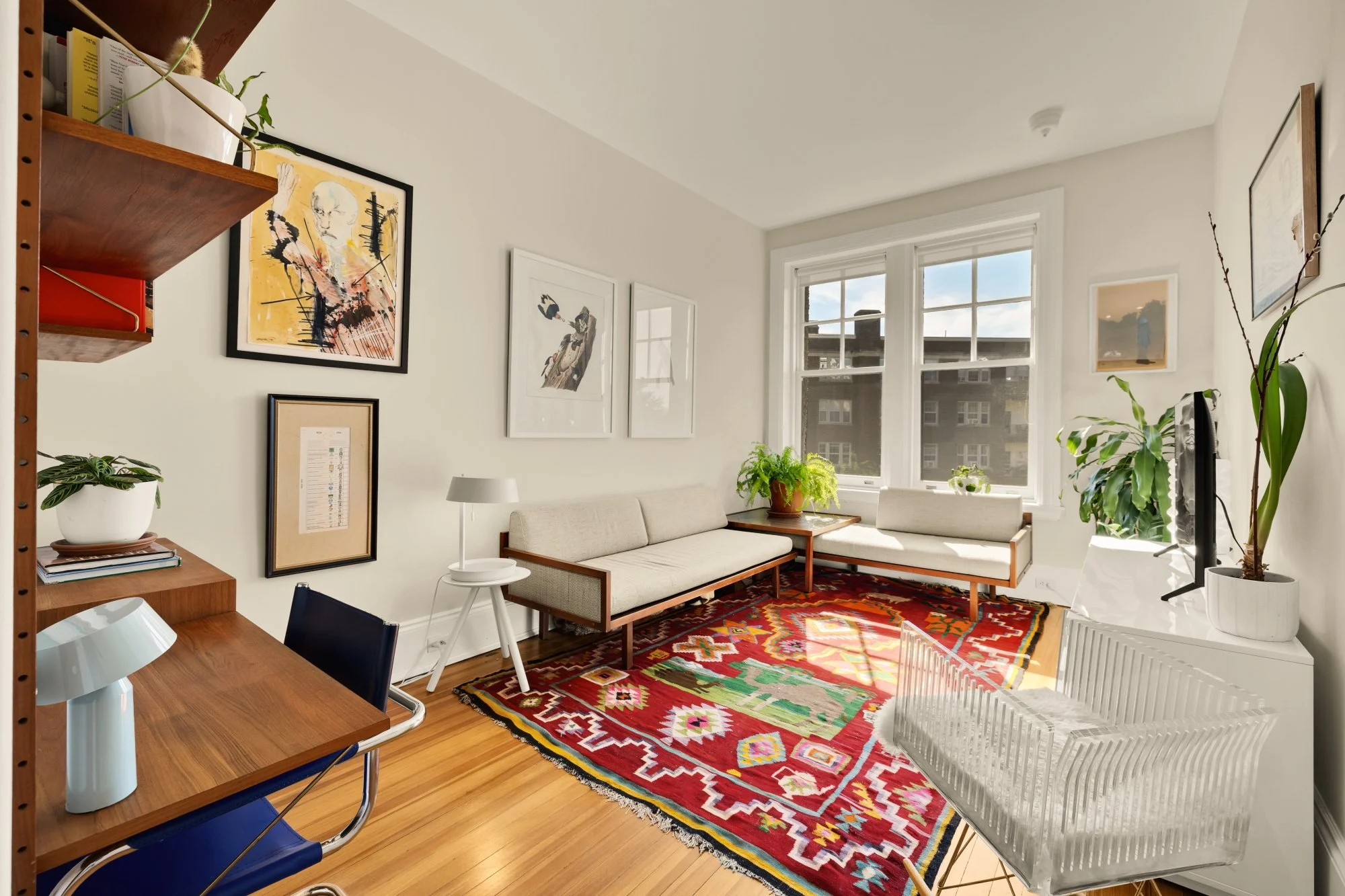 Bright living room with white walls and large windows, furnished with beige sofas, potted plants, a red patterned area rug, and wall art.