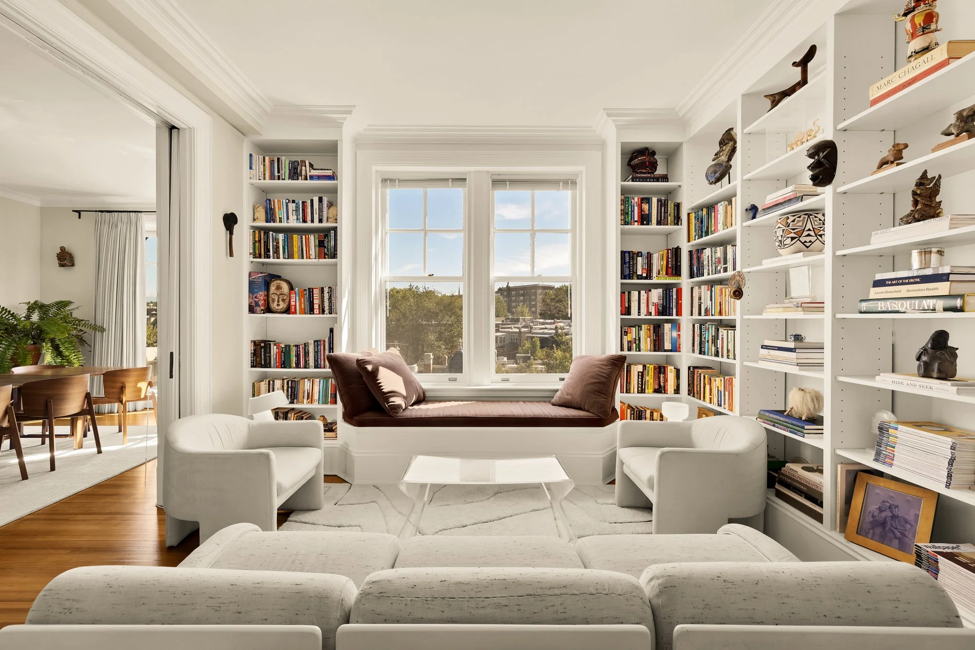 Bright living room with large window, white bookshelves filled with books and decorations, white armchairs, and a window seat with brown cushions.
