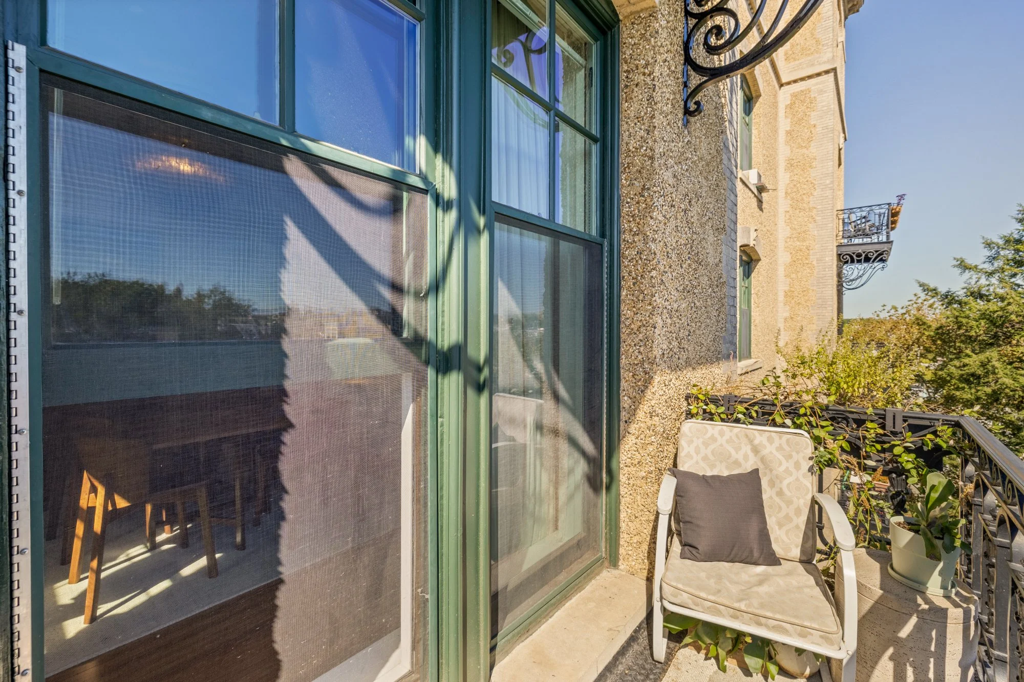 A small balcony with a beige patterned chair and a black pillow, potted plants, a decorative black iron railing, adjacent windows with green frames, and part of a building with beige textured exterior, under a clear blue sky.