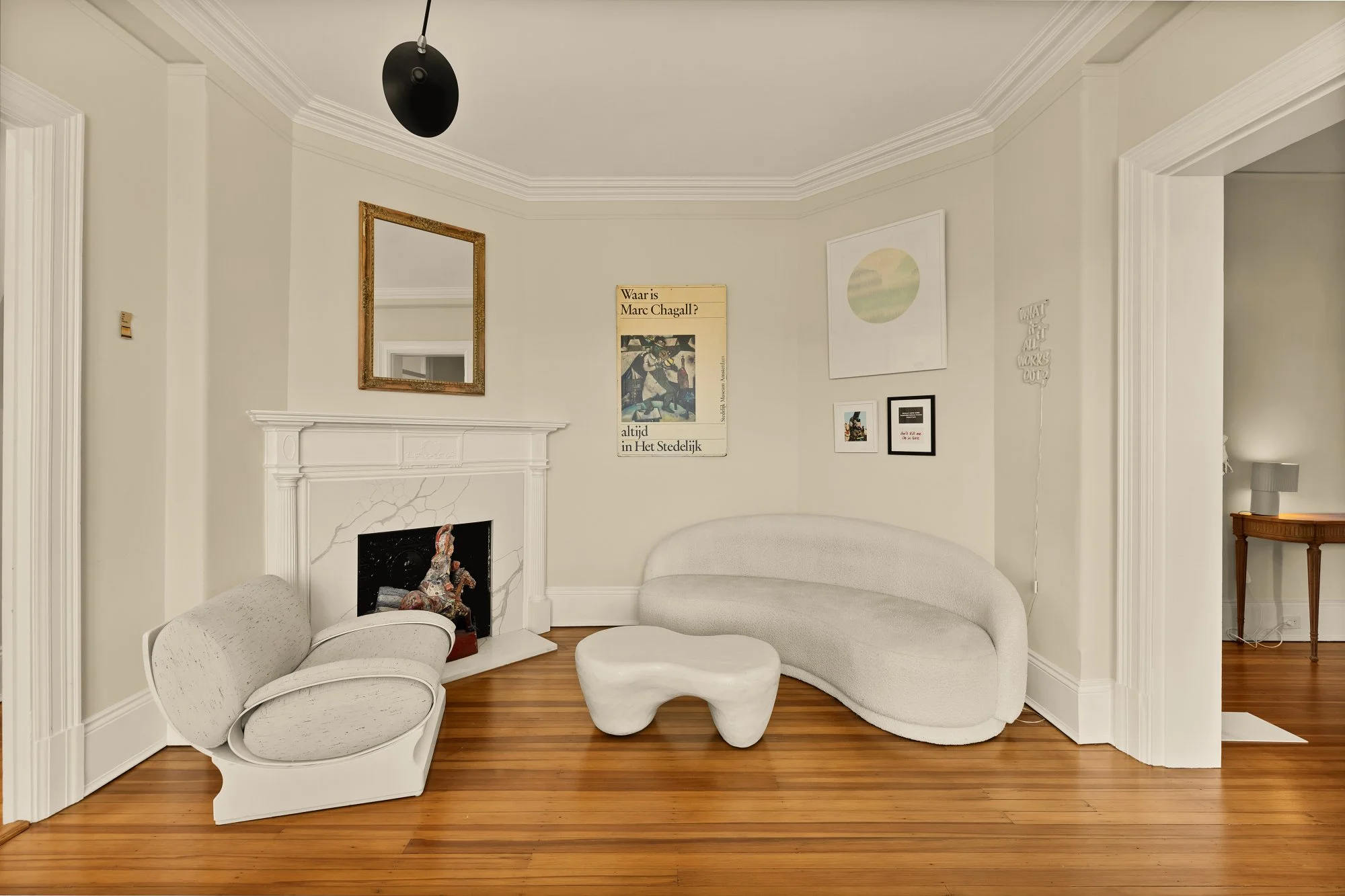 Living room with a white curved sofa, a unique white ottoman, a vintage armchair with a white cushion, a fireplace with a decorative piece inside, and wall art including a mirror, posters, and framed pictures. Hardwood floor and cream walls with crow