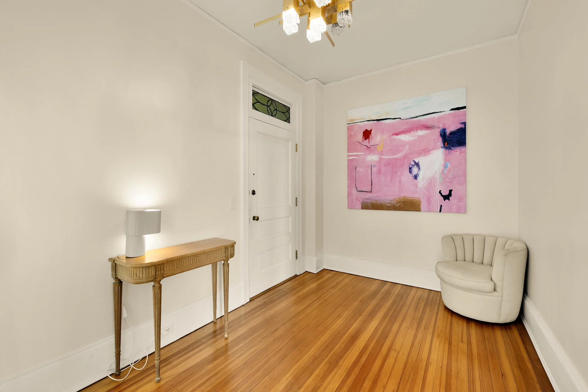 An empty room with a floral chandelier, white walls, a wooden floor, a small beige corner chair, a narrow wooden side table with a white lamp, a white door, a small window above the door, and a large colorful abstract painting on the wall.