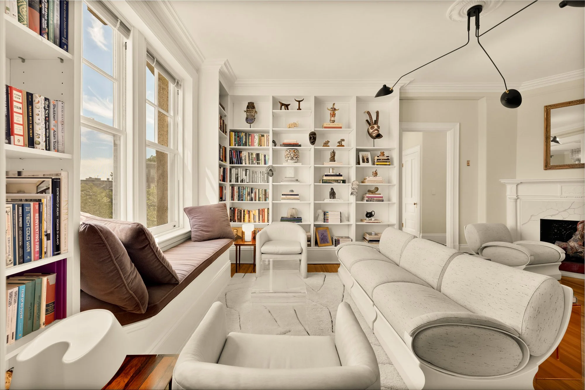 Living room with large windows, white bookshelf with decorative masks and books, cream-colored sofa, armchair, window seat with pillows, and a fireplace with a mirror above it, in a bright and modern interior.