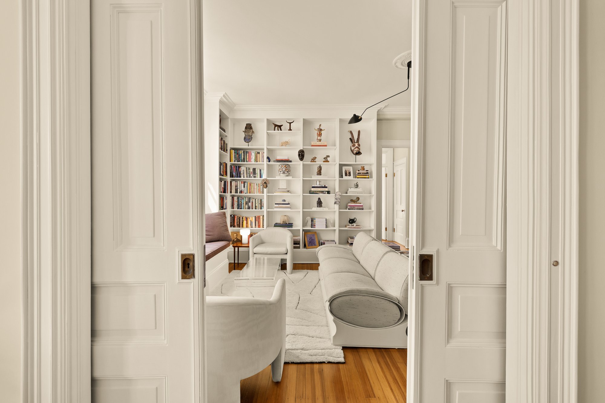 Living room with white built-in bookshelves, various decorative items, and a seating area with white and beige furniture, hardwood floors, and a sliding door opening into the room.