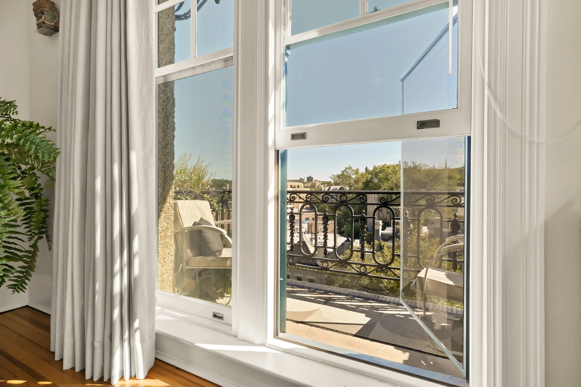View through an open balcony door showing wrought iron railing, outdoor chairs, and trees in the distance during daytime.