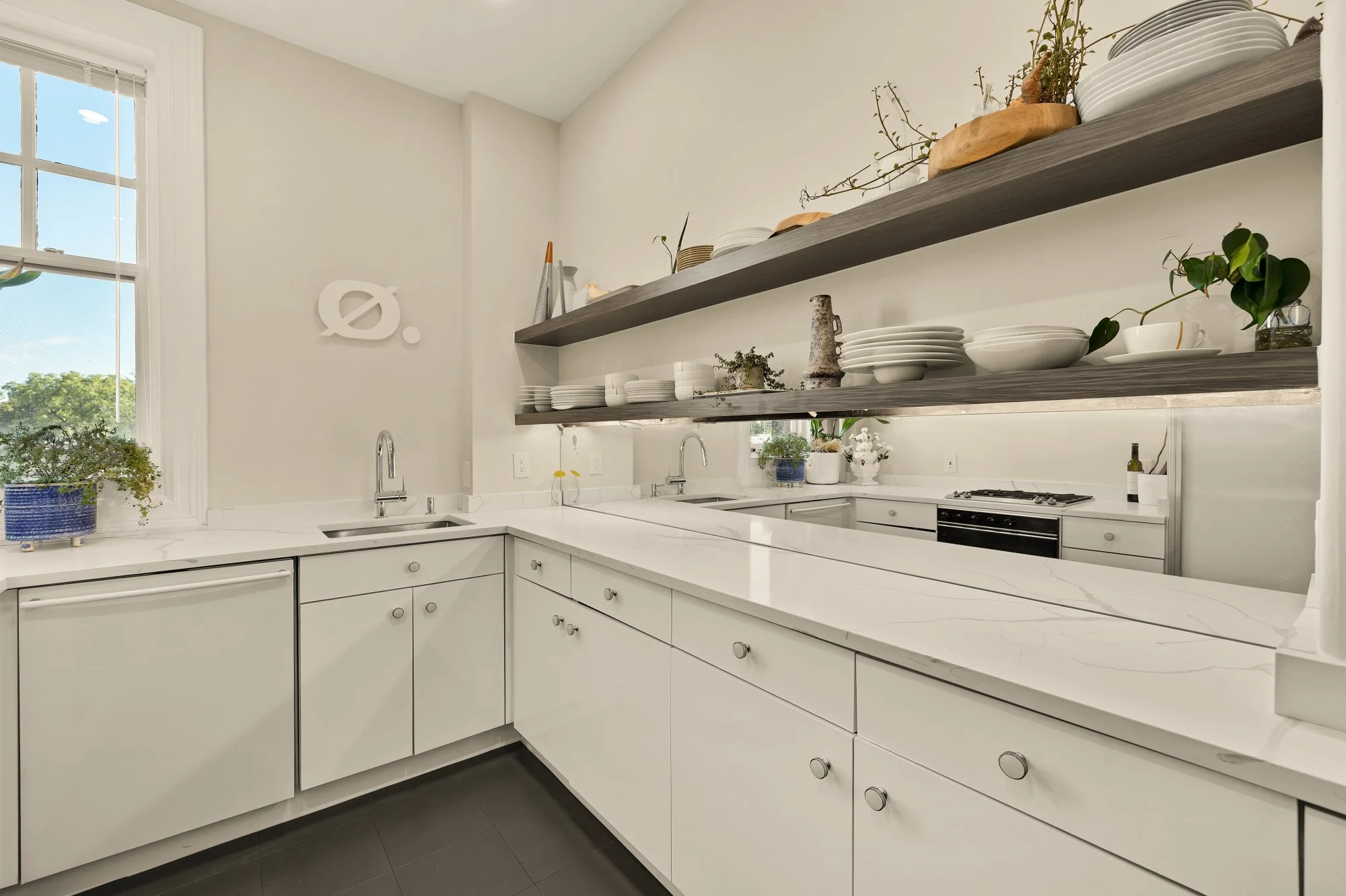 Modern kitchen with white cabinets, marble countertops, open grey shelves with dishes and plants, and a large window.