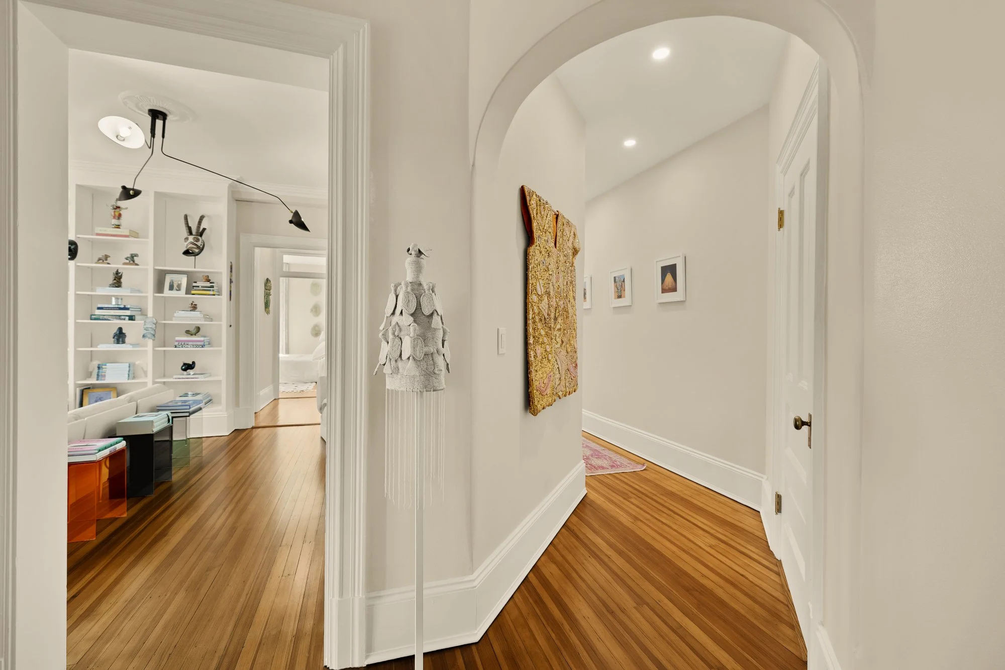 Interior view of a hallway with hardwood floors, white walls, and art decor. There is an archway leading to another room and a separate doorway to the right. A colorful woven wall hanging is on the wall, and framed pictures line the hallway. Bright l
