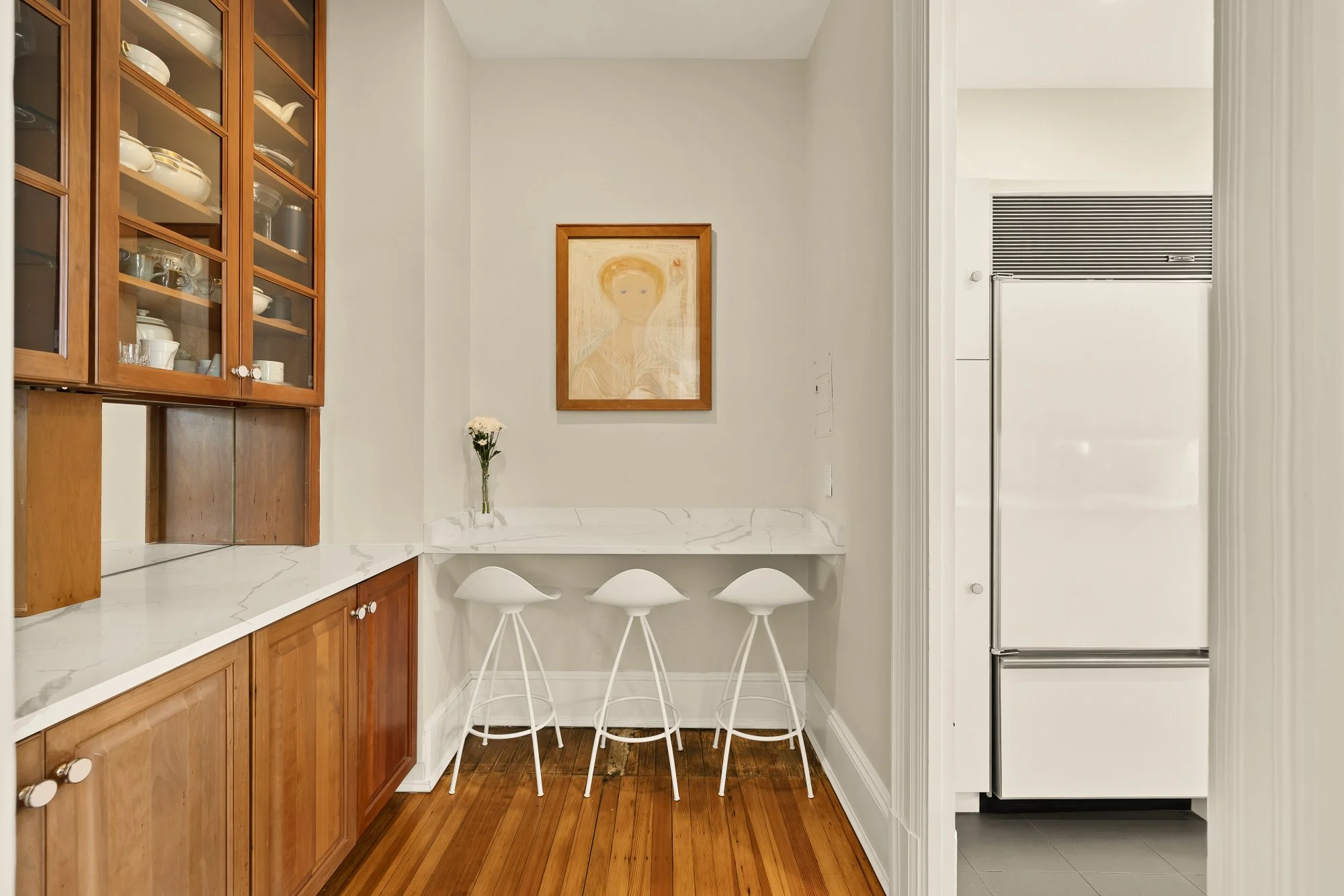 A small dining area with a marble-top table, three white stools, a modern art painting on the wall, a flower in a glass vase, and wooden kitchen cabinets to the left. An open doorway reveals a modern white refrigerator inside a kitchen.