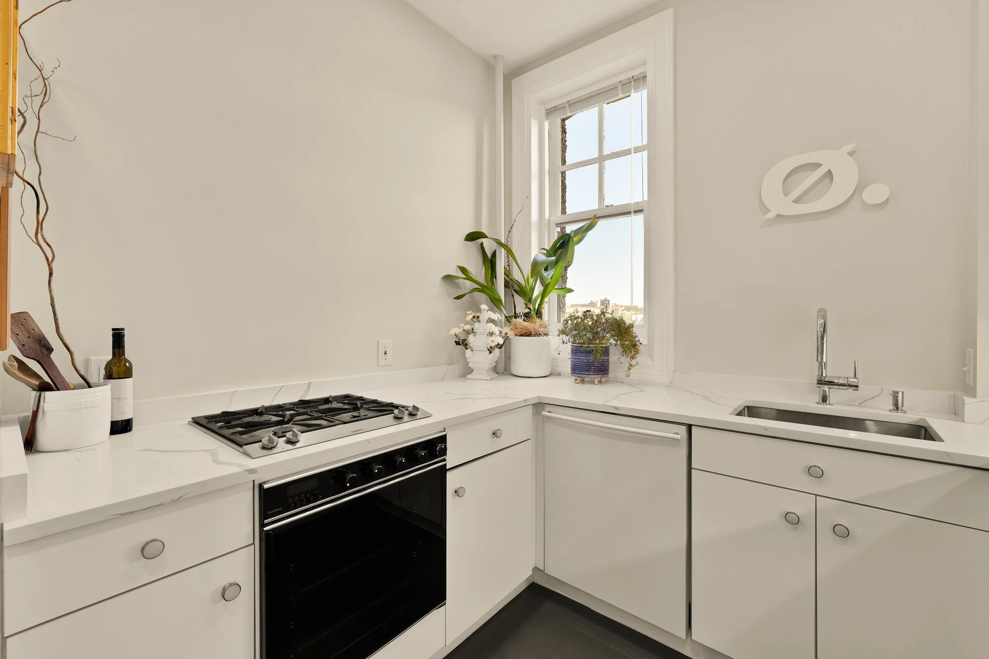A white kitchen with a marble countertop, a black oven stove, and white cabinets. There are three potted plants on the counter by a window; two have green leaves, one has small white flowers, and the other has brownish-green foliage. On the wall, the