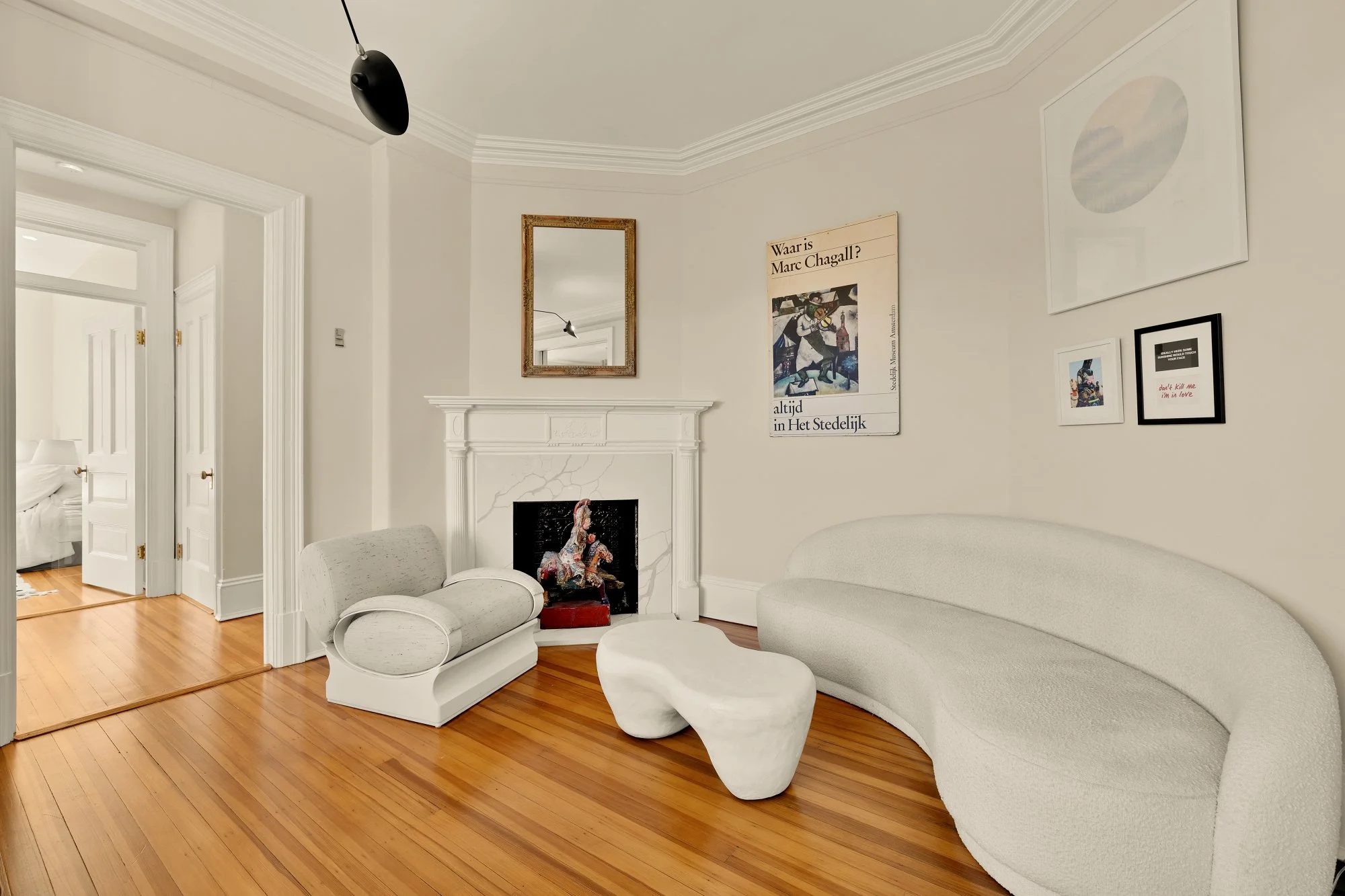 Living room with white modern curved sofa, white rocking chair, and abstract shaped white ottoman. Artworks on the wall, including a large framed print, smaller frames, and a mirror above the white fireplace. Hardwood floors and a doorway leading to 