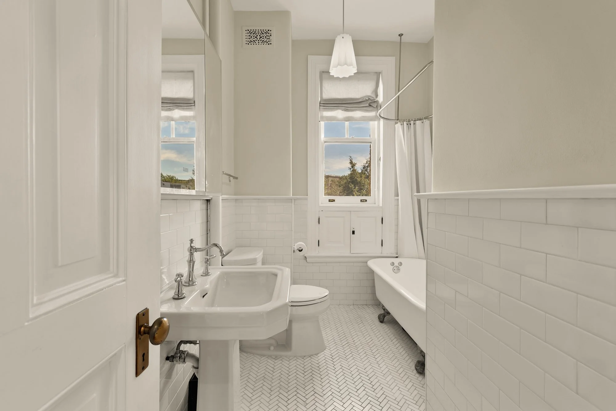 A vintage bathroom with a clawfoot bathtub, a toilet, a pedestal sink, white subway tiles, a window with a view of trees, a hanging lampshade, and a shower curtain.