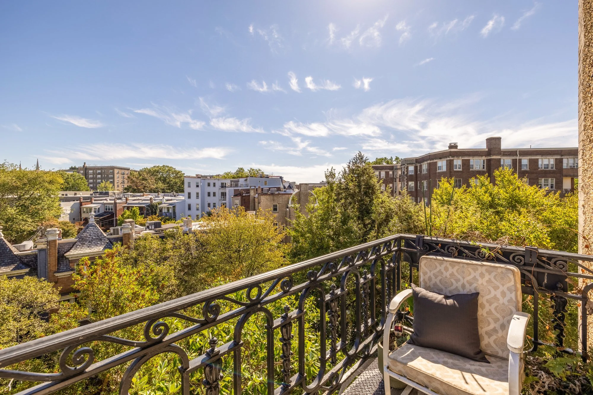 View from a balcony overlooking trees and city buildings under a partly cloudy sky, with a cushioned chair on the balcony.