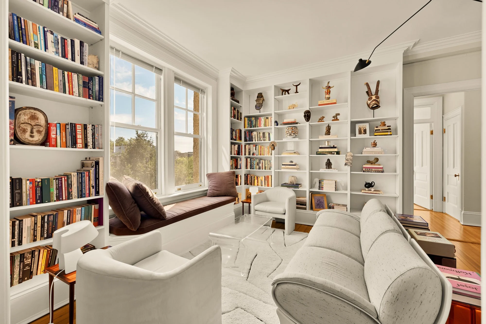 Bright living room with white walls, large windows, and built-in white bookshelves filled with books and decorative items. There is a white sofa, a white armchair, and a window seat with brown cushions. A glass coffee table and a cream-colored rug ar