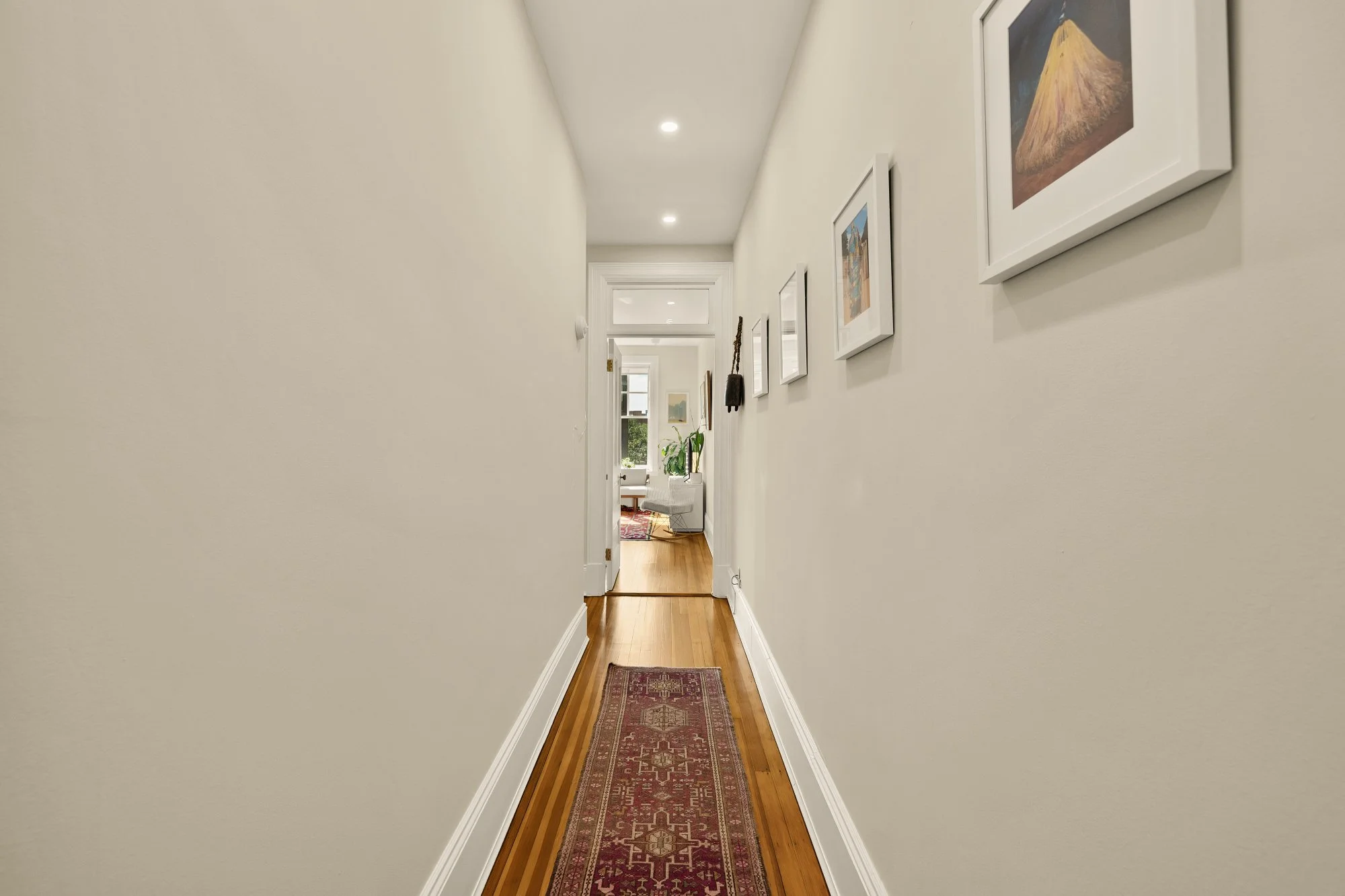 A narrow hallway in a house with white walls, wooden flooring, and framed artwork hanging on the right wall leading to a bright living room area.
