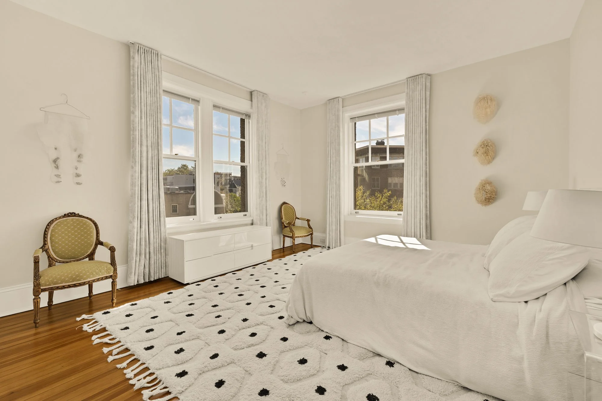 Bright bedroom with white walls, hardwood flooring, and a large white bed. Natural light streams through three windows with white curtains. There are two vintage-style chairs, one near the windows and one against the wall, and a white dresser. Decor 