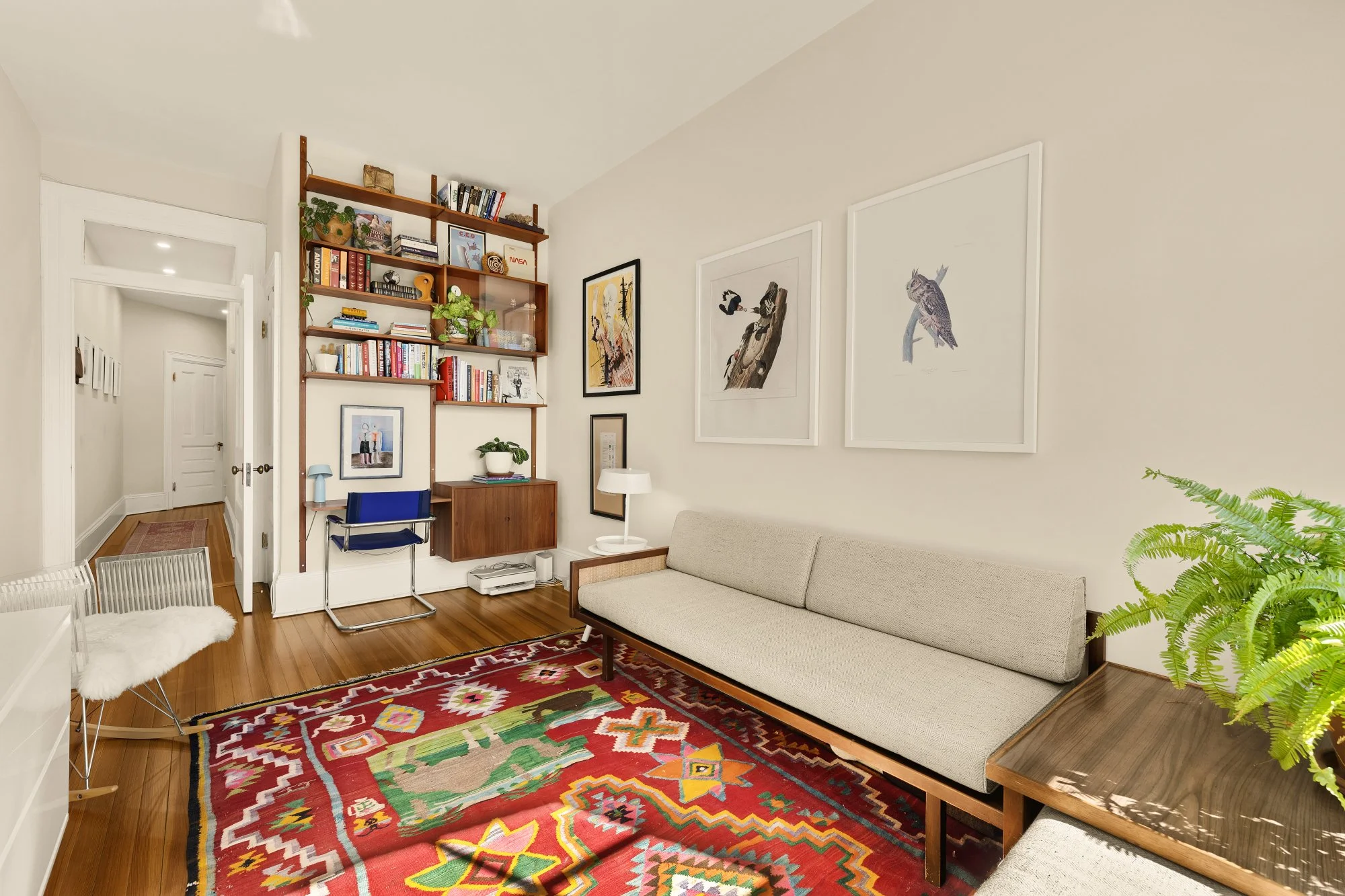 Living room with a beige sofa, a colorful patterned rug, a wooden side table with a green plant, a white lamp, and a floor-to-ceiling bookshelf filled with books and decorative items. There are framed artworks on the wall above the sofa.