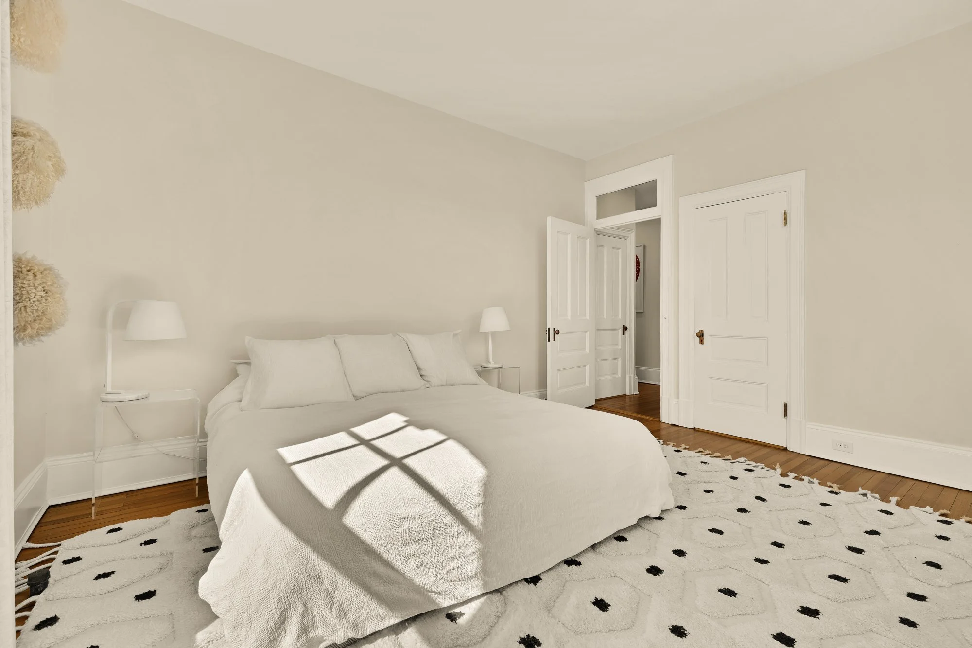Bedroom with a white bed, white walls, a nightstand with a white lamp, a white rug with black patterns, and doors leading to other parts of the house.