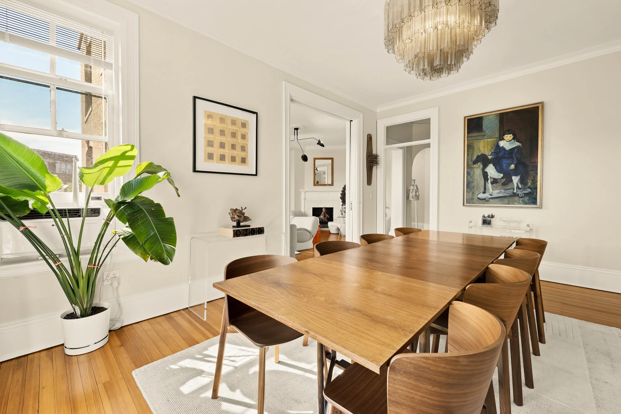 A dining room with a wooden table and eight matching wooden chairs. There is a large window on the left, a potted plant near it, and framed artwork on the walls. The room has hardwood floors and white walls with crown molding.
