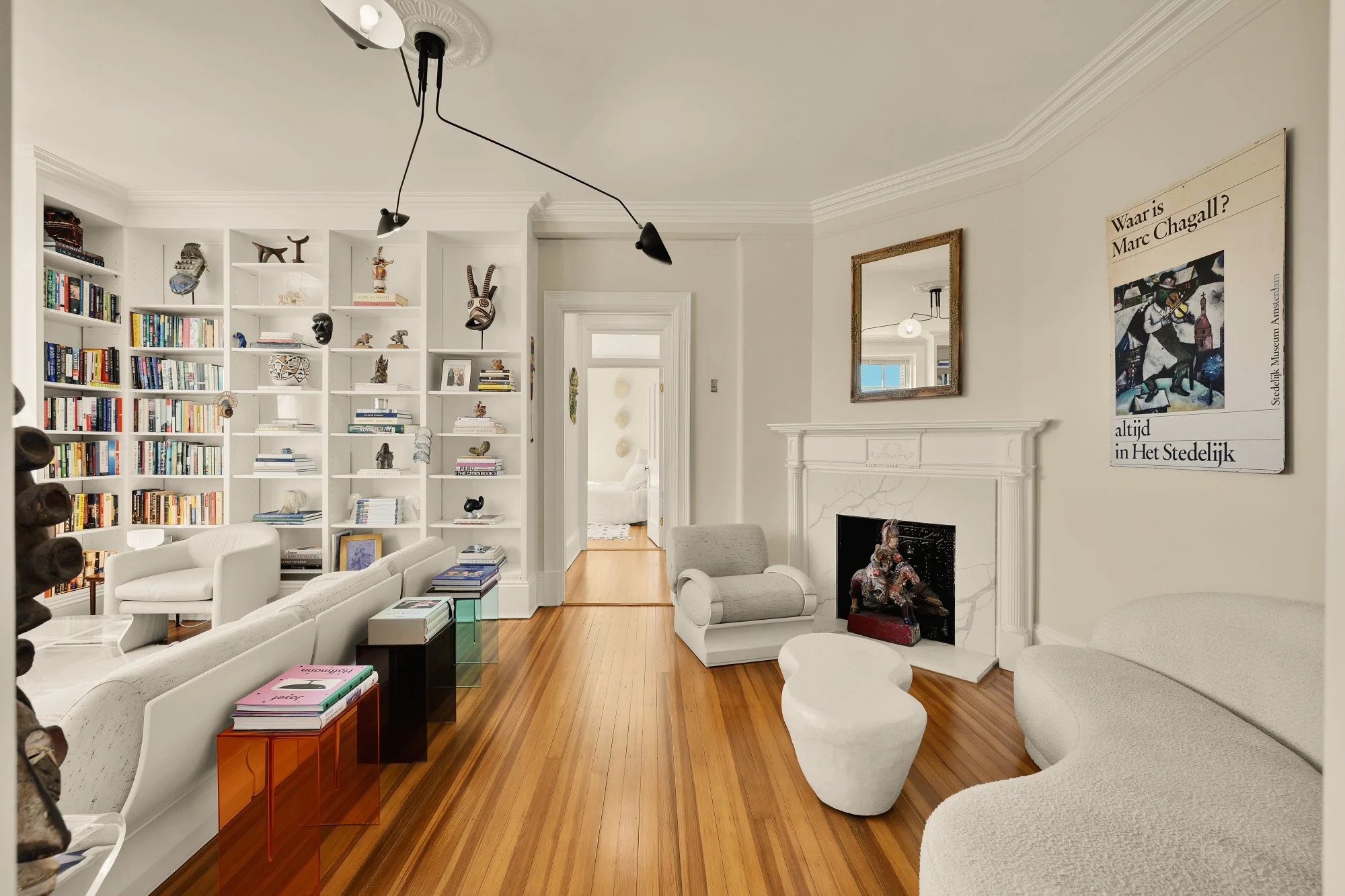 Living room with white walls, hardwood floors, a white bookshelf filled with books and decorative items, a white couch, and modern white chairs. A fireplace with a mirror above it and a large artwork on the wall. The room has contemporary furniture a