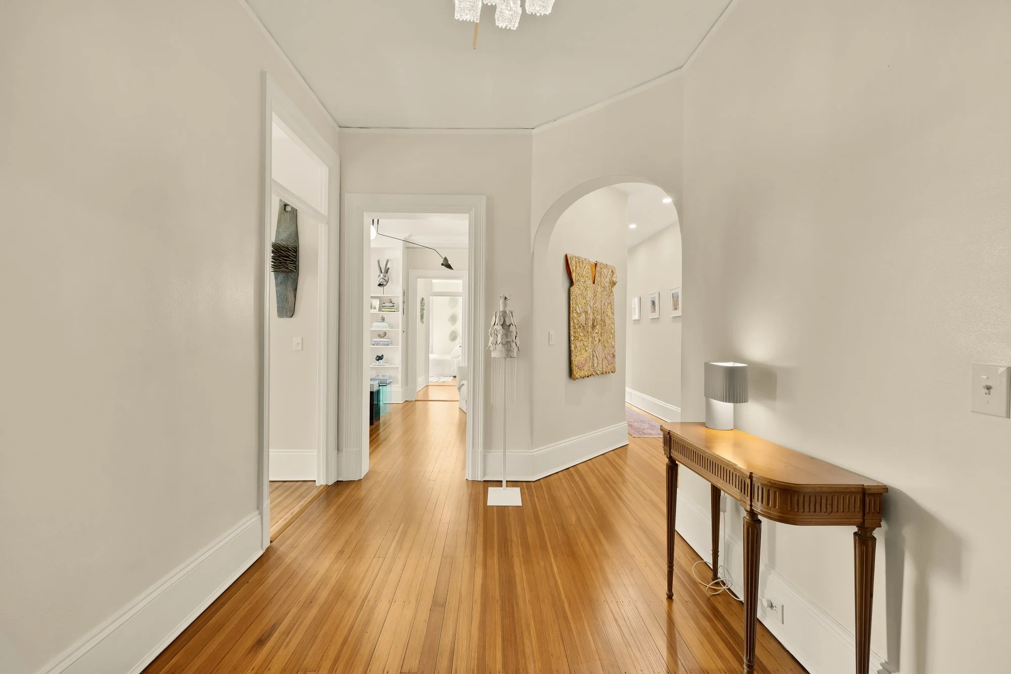 A hallway in a home with wooden flooring, white walls, and simple decor including a table with a lamp and wall art.