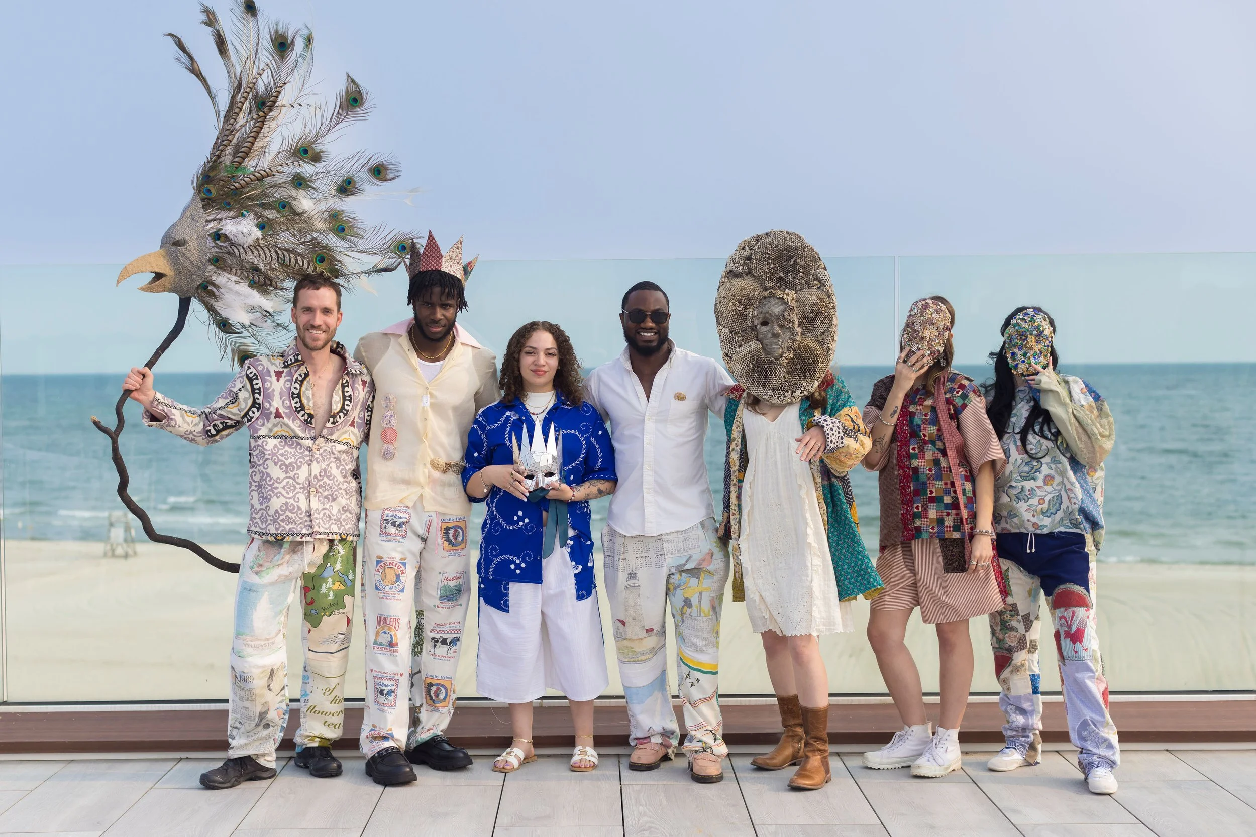 Arts Gala, Arts Foundation of Cape Cod, Group of seven people dressed in colorful, eclectic clothing with some wearing masks and headpieces, standing on a boardwalk by the beach with ocean in the background.