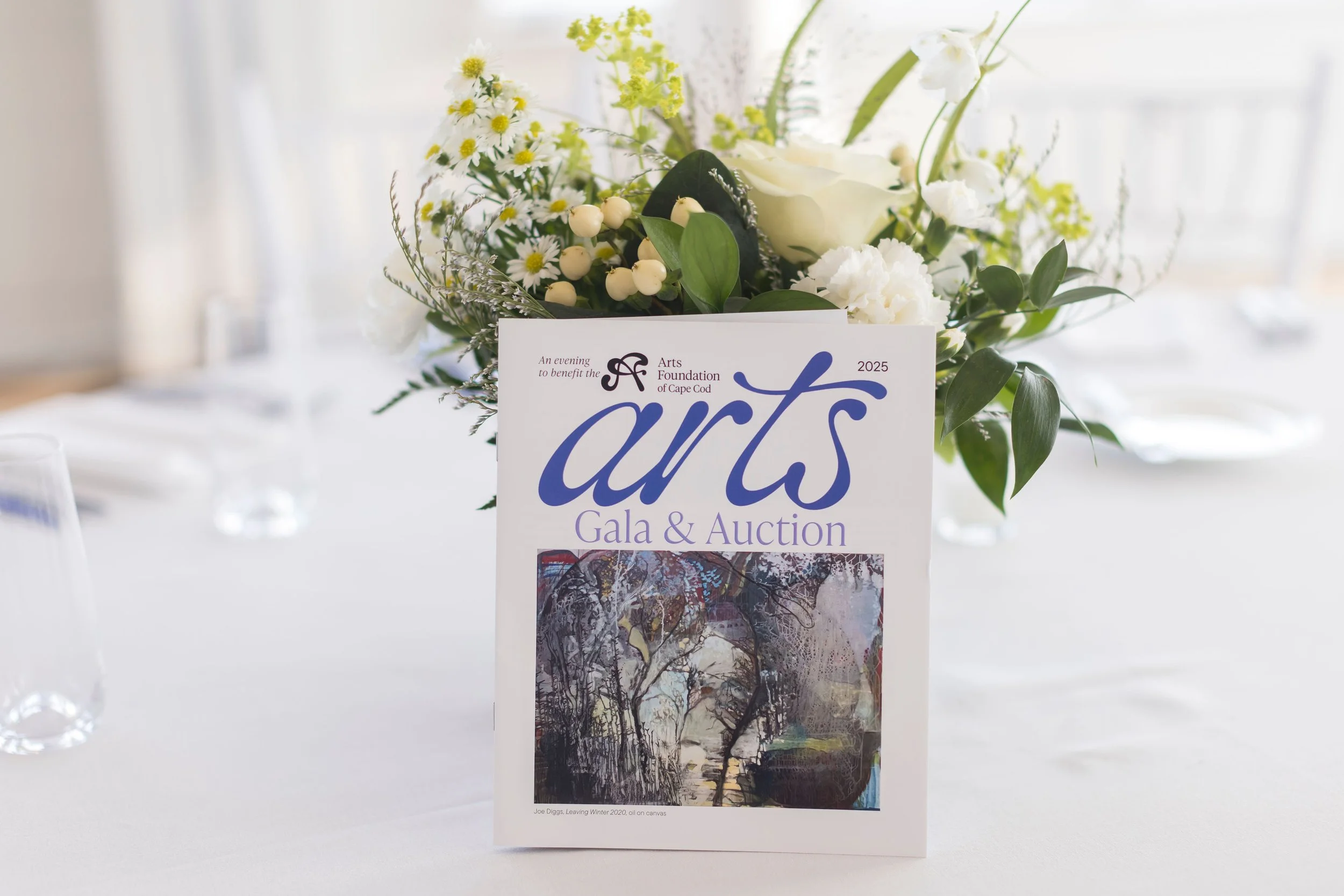 Arts Gala, Arts Foundation of Cape Cod, A floral centerpiece and an art foundation event program on a white table, with clear glasses in the background.