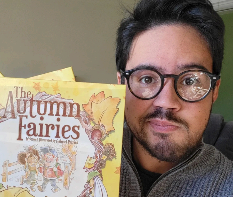 A man holding a children's book titled 'The Autumn Fairies' with an illustrated cover of two fairies and autumn leaves, in a room with a green wall.