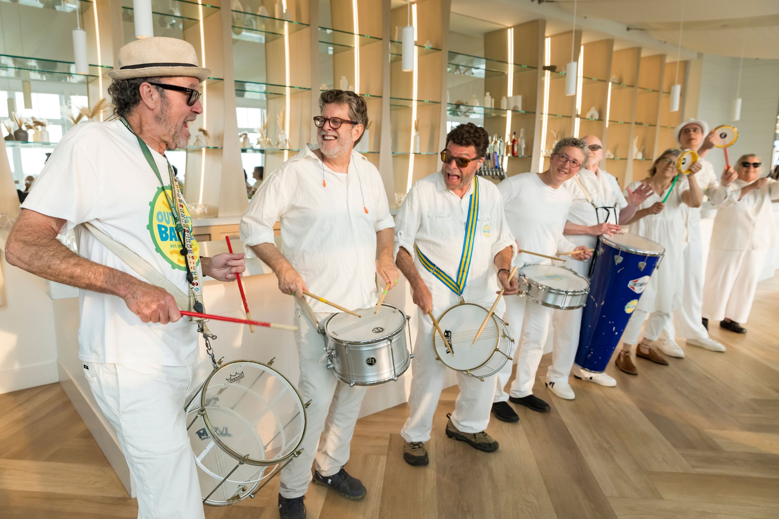 Group of seniors dressed in white, playing drums and percussion instruments together in a brightly lit, modern indoor space.