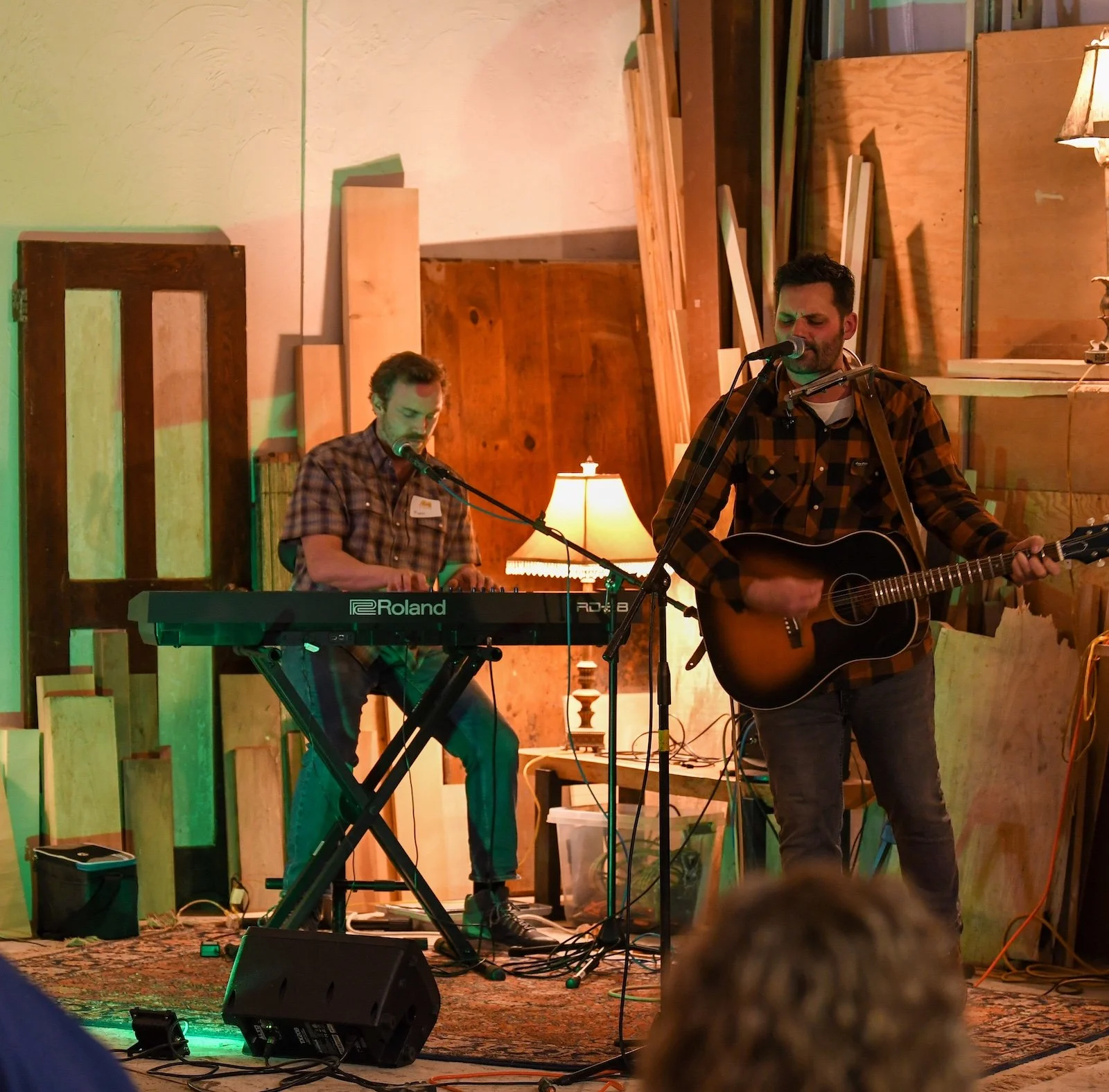 Two musicians performing in a cozy indoor setting, one playing keyboard and the other playing guitar and singing, surrounded by wood panels and warm ambient lighting.
